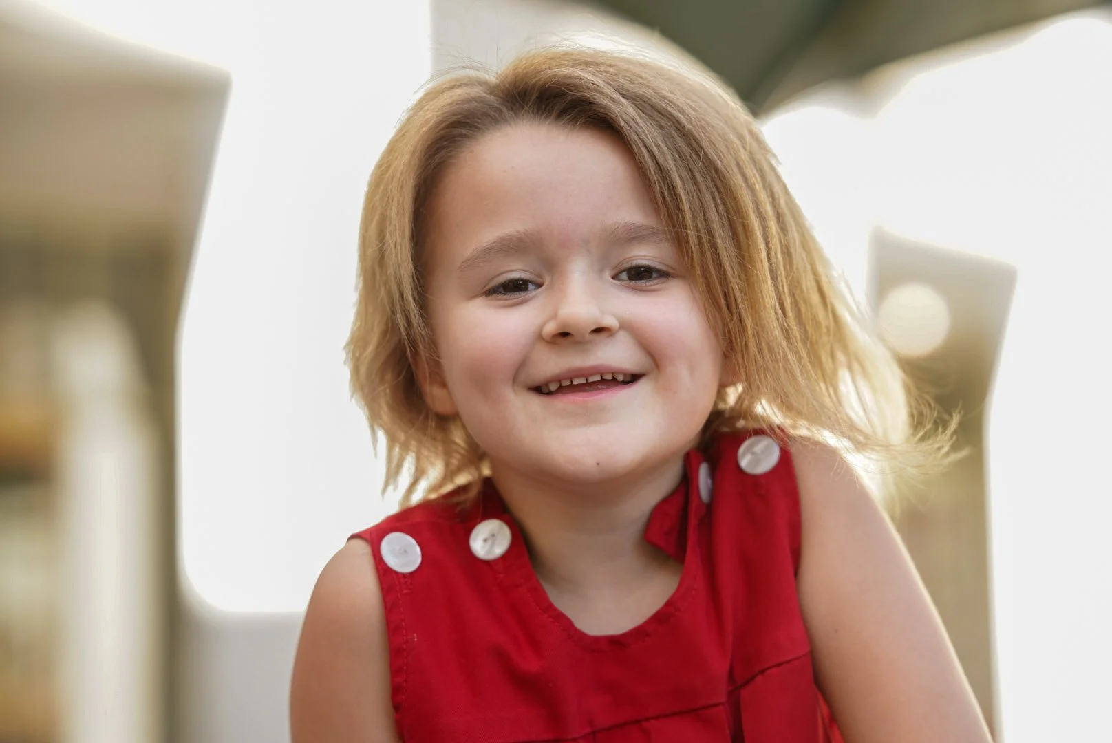A young girl with light brown, shoulder-length hair smiling, wearing a sleeveless red top with white buttons on the shoulders.