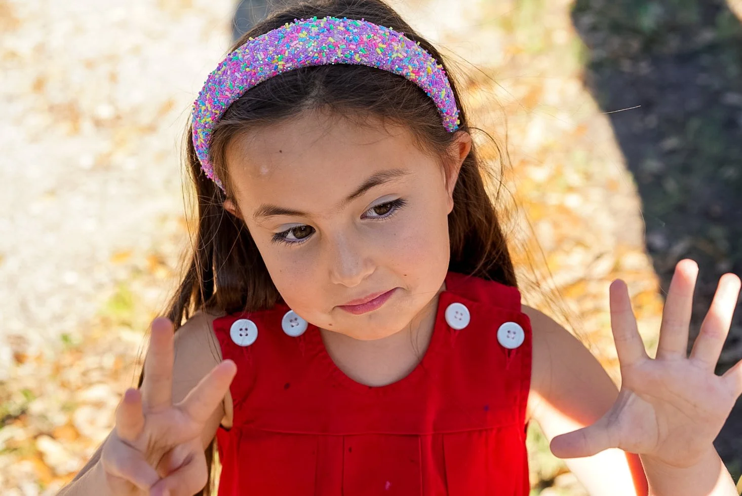 A young girl with long brown hair, wearing a red dress with white buttons and a colorful sparkly headband, making peace signs with both hands outdoors.