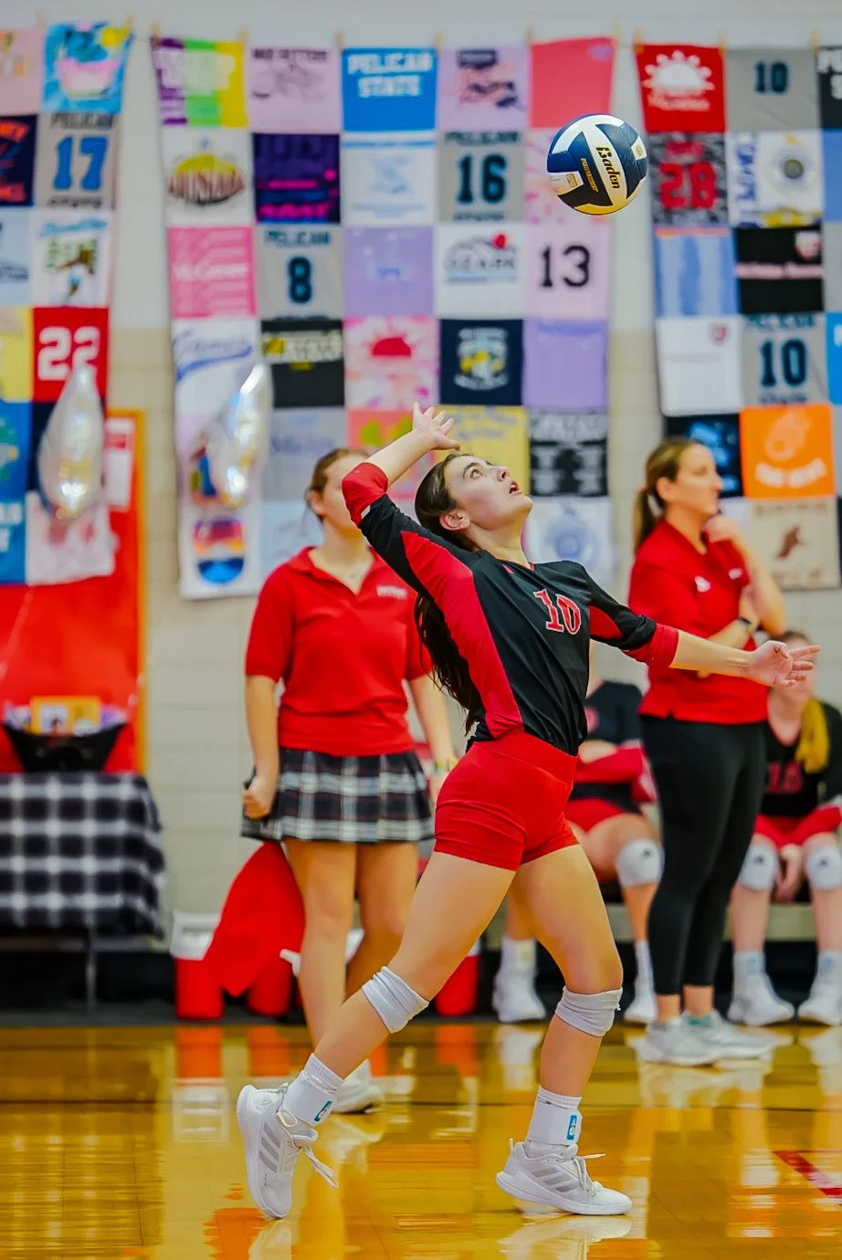 A female volleyball player wearing a black and red uniform with the number 10 prepares to hit a volleyball during a game. She is on a gymnasium court, with her teammates and a colorful banner with various numbers hanging in the background.