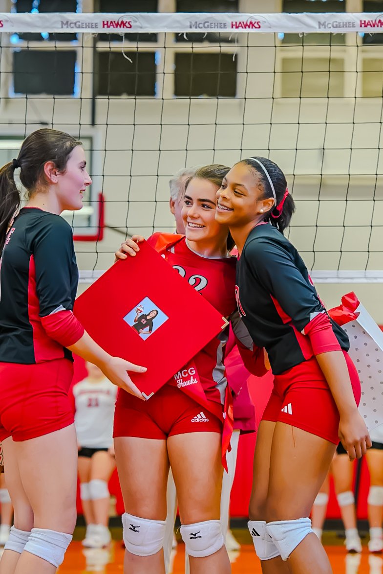 Three young female volleyball players in red and black uniforms smiling and celebrating on an indoor volleyball court. One girl is holding a red box with a printed image on it.
