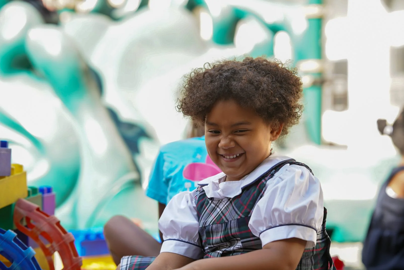 Young girl with curly hair smiling while playing with colorful building blocks in a classroom.