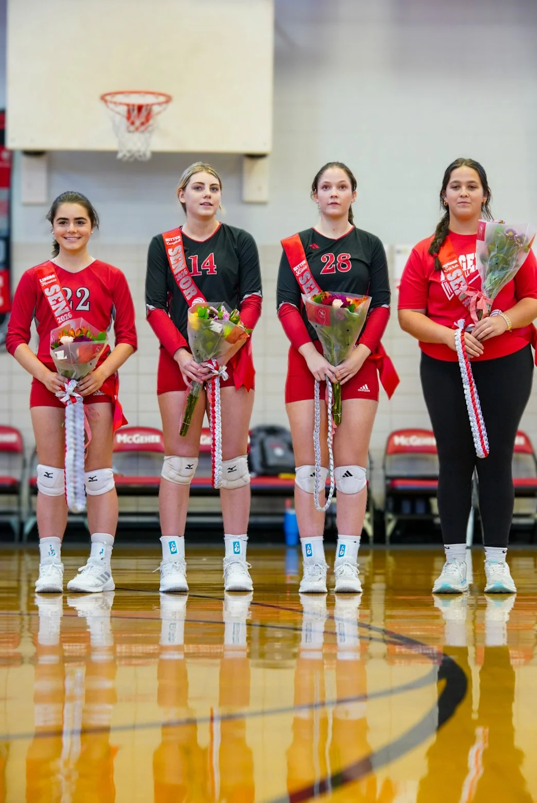 Four female volleyball players standing on a gymnasium court, holding bouquets of flowers, wearing medals, with a basketball hoop above them.
