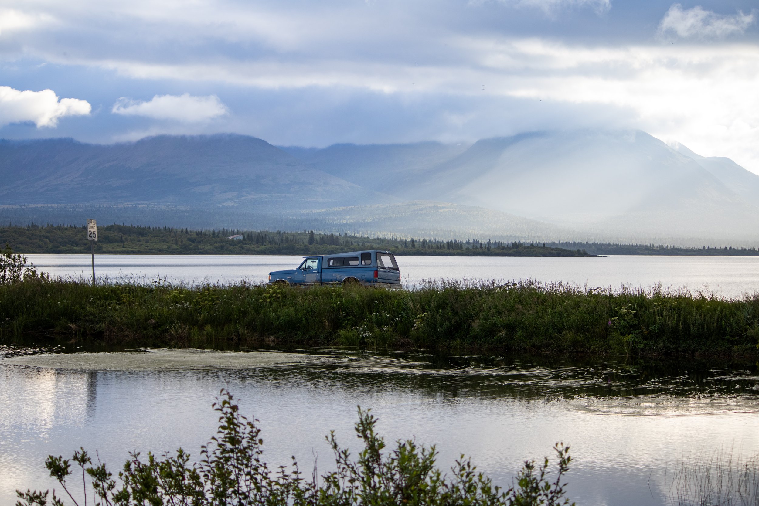 A blue vehicle parked on a grassy area near a body of water with mountains and cloudy sky in the background.