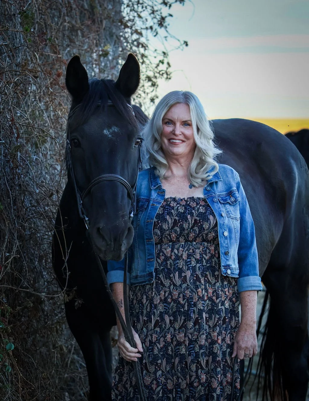 A woman with blonde hair wearing a denim jacket and floral dress standing next to a black horse outdoors at sunset.