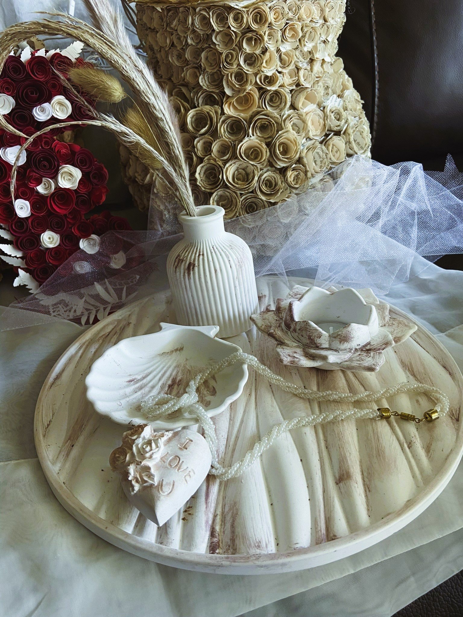 Decorative arrangement with white and beige ceramic dishes, a small white vase with dried grasses, a pearl necklace, and a heart-shaped ornament with the words 'I love you' on a beige tray, with a background of cream and red roses.