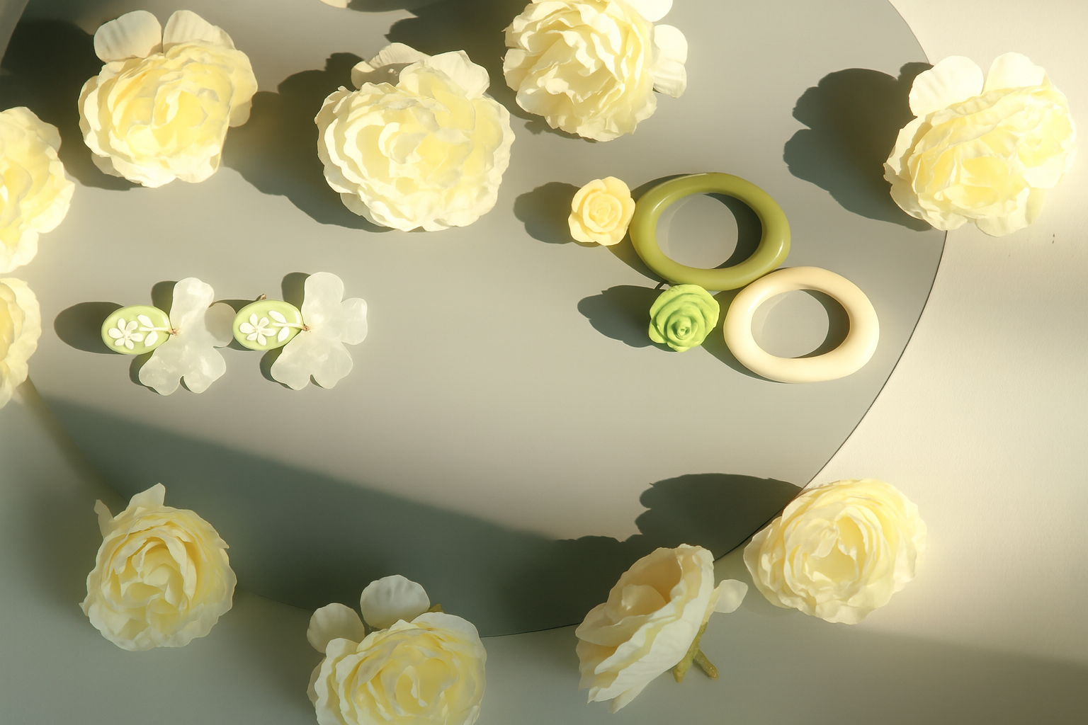 Cream-colored roses and white flowers arranged around a round mirror with green and white rings and small flowers on top, with shadows cast by sunlight.