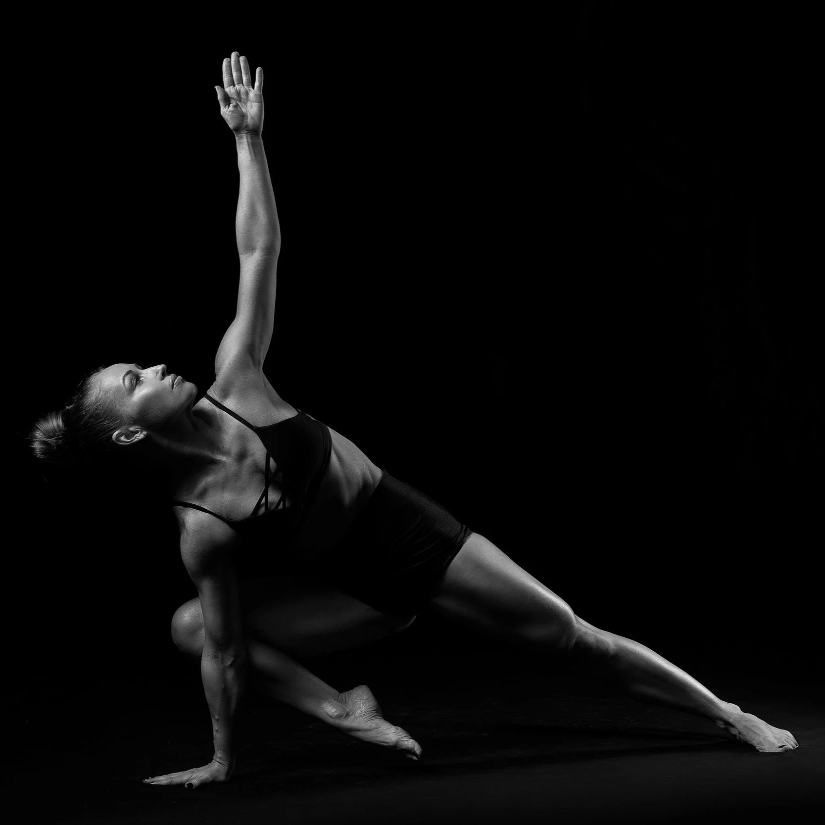 Black and white photo of a woman practicing yoga, balancing on one hand and one foot, with the other arm extended upward.
