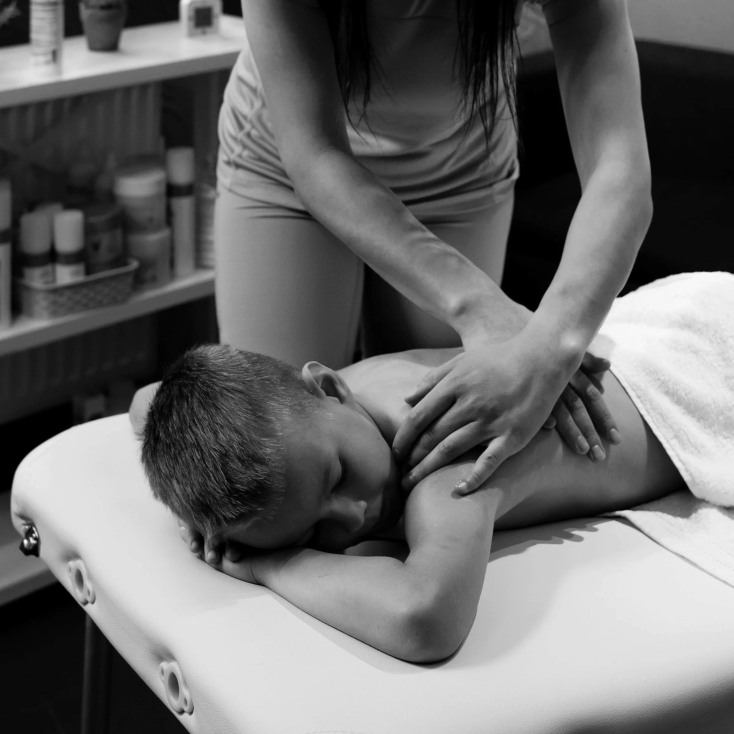 A young boy is lying face down on a massage table, receiving a back massage from a therapist in a room with shelves of skincare products.