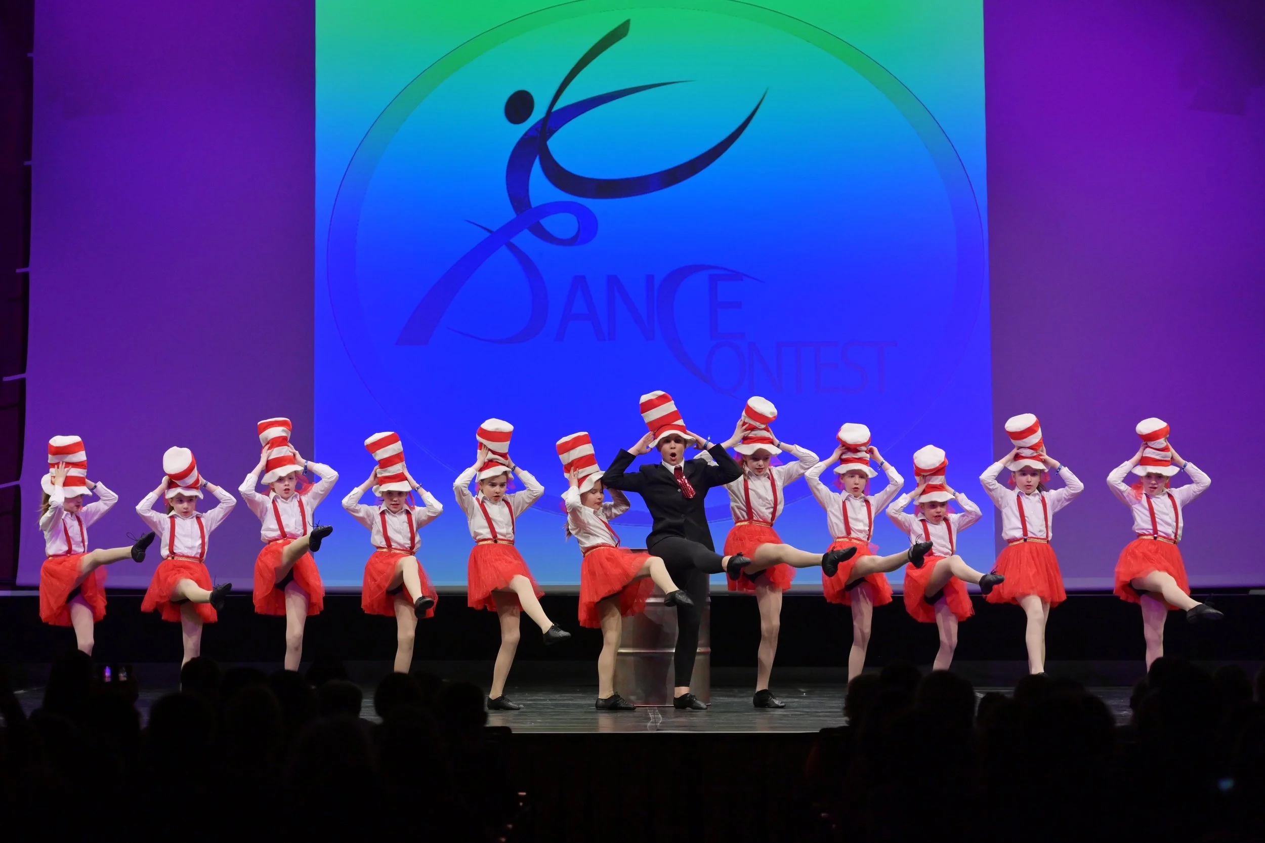 Tanzgruppe auf der Bühne mit roten Röcken, weißen Hemden, roten Hosenträgern und rot-weiß gestreiften Hüten bei einem Tanzwettbewerb, im Hintergrund ein Bildschirm mit dem Text 'Dance Contest'.