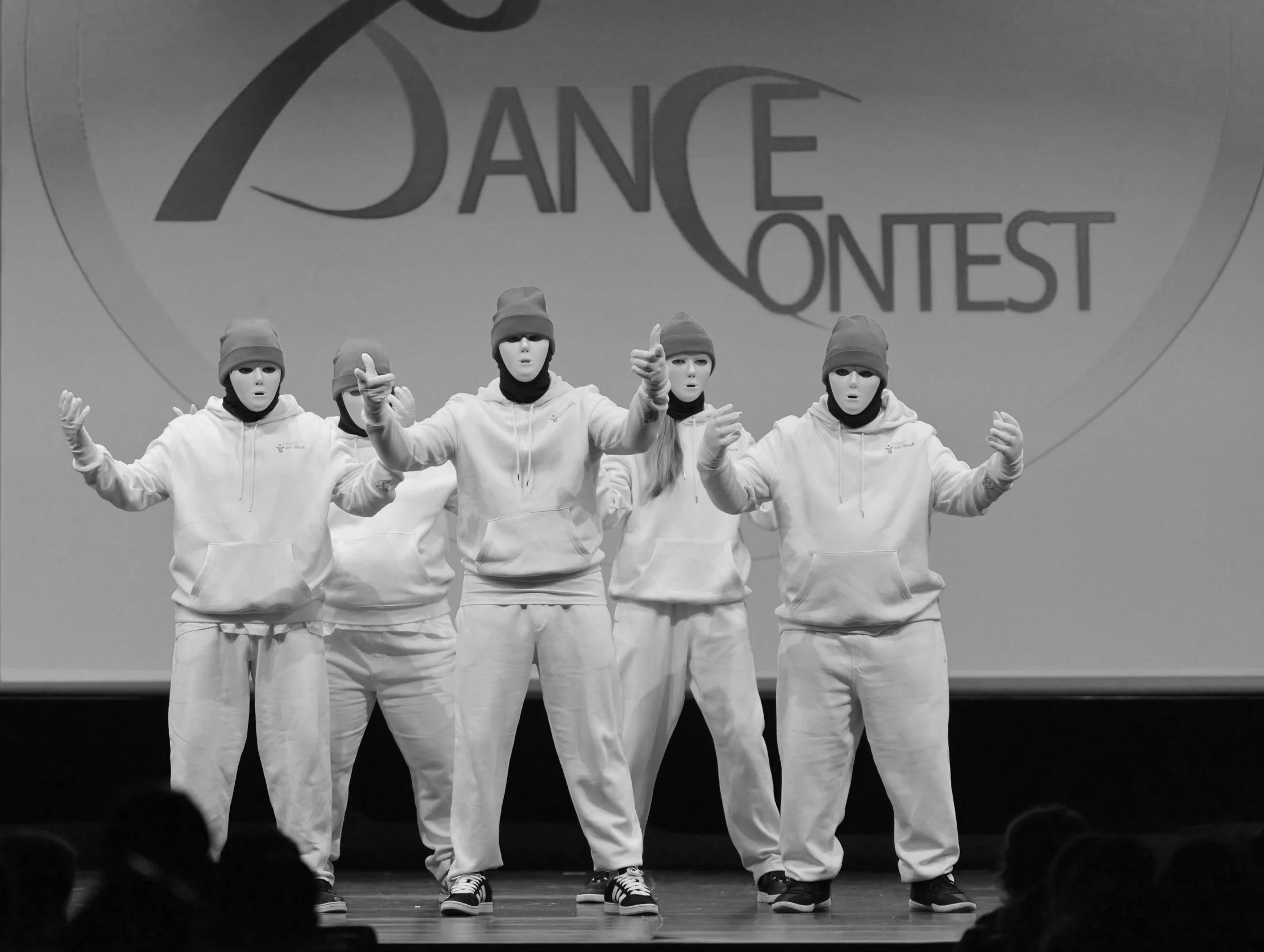 Tanzgruppe in weißen Hoodie, Jogginghosen und Masken während eines Tanzwettbewerbs auf der Bühne, mit dem Hintergrundtext 'Dance Contest'.