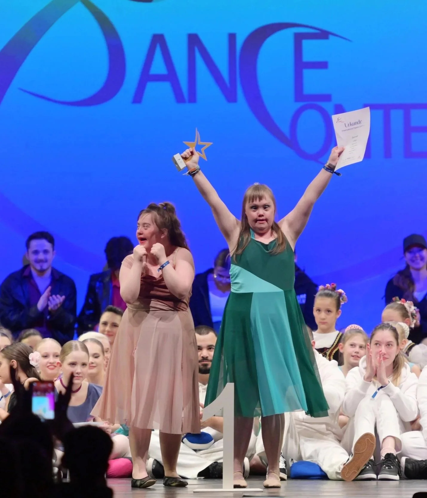 Siegerin mit Statuette und Urkunde auf Bühne bei Tanzwettbewerb, umgeben von lachenden und applaudierenden Menschen, im Hintergrund großes Logo 'Dance'.