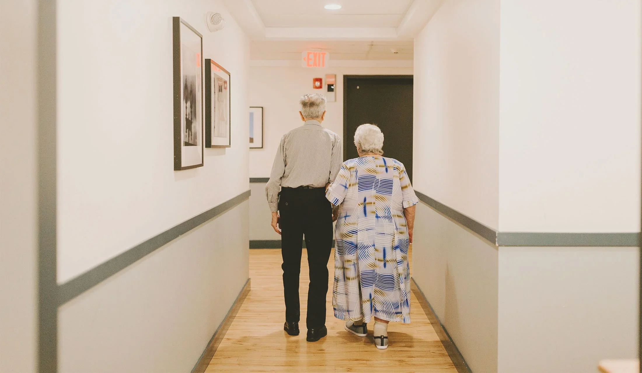 An elderly woman and a younger person walking down a hallway with white walls and framed pictures, with an exit sign visible at the end of the hallway.