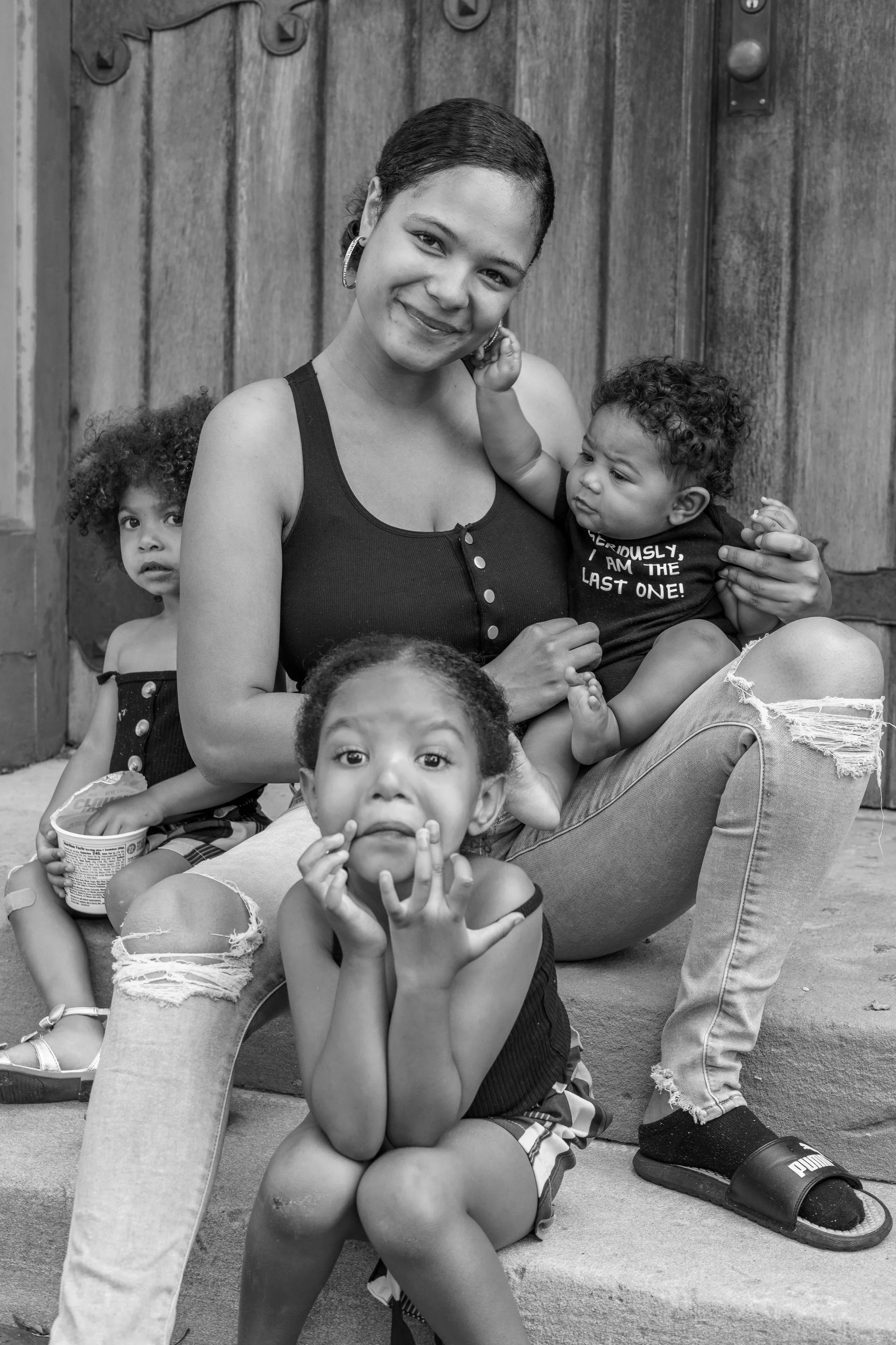 A woman sitting outdoors on steps, surrounded by four young girls, with a wooden fence in the background. The woman is smiling, wearing a black tank top and ripped jeans. The girls are making various facial expressions and are casually dressed.