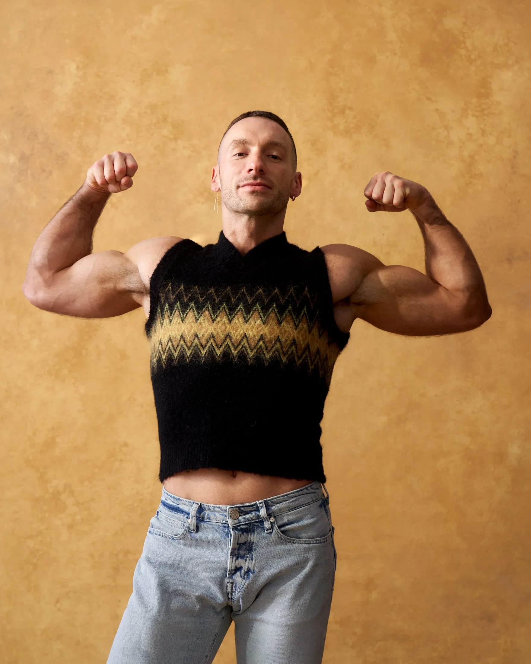 A man flexing his biceps, wearing a sleeveless black sweater with a zigzag pattern and light blue jeans, against a yellow textured background.