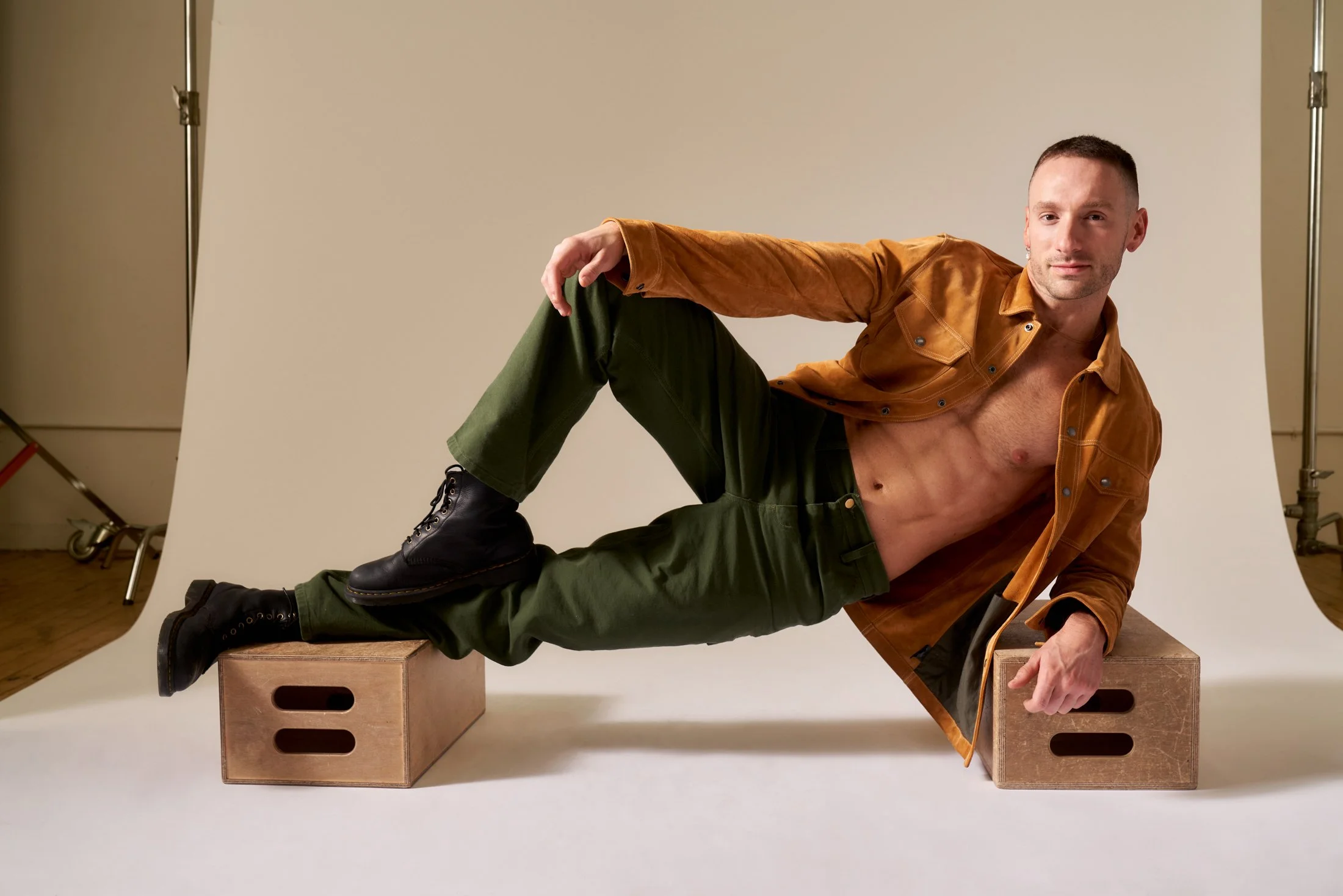 A shirtless man wearing a brown jacket, green pants, and black boots lying sideways on two wooden boxes in a studio with a neutral background.