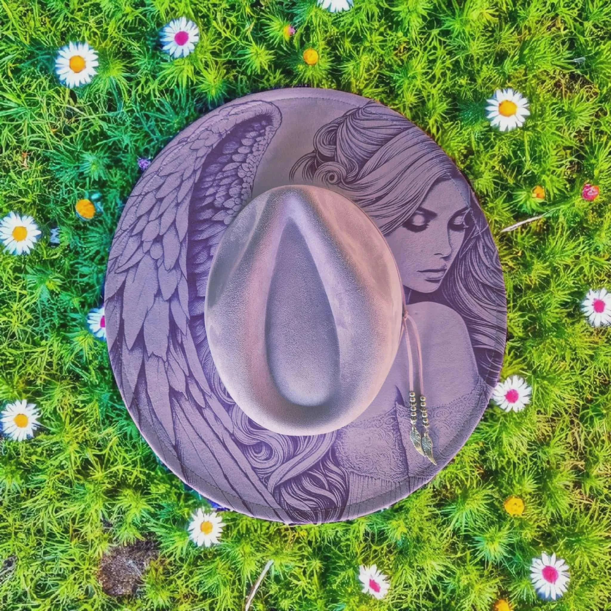 Top view of a round hat with a sketch of a woman with long hair and winged creature, placed on green grass dotted with white daisies and yellow flowers.