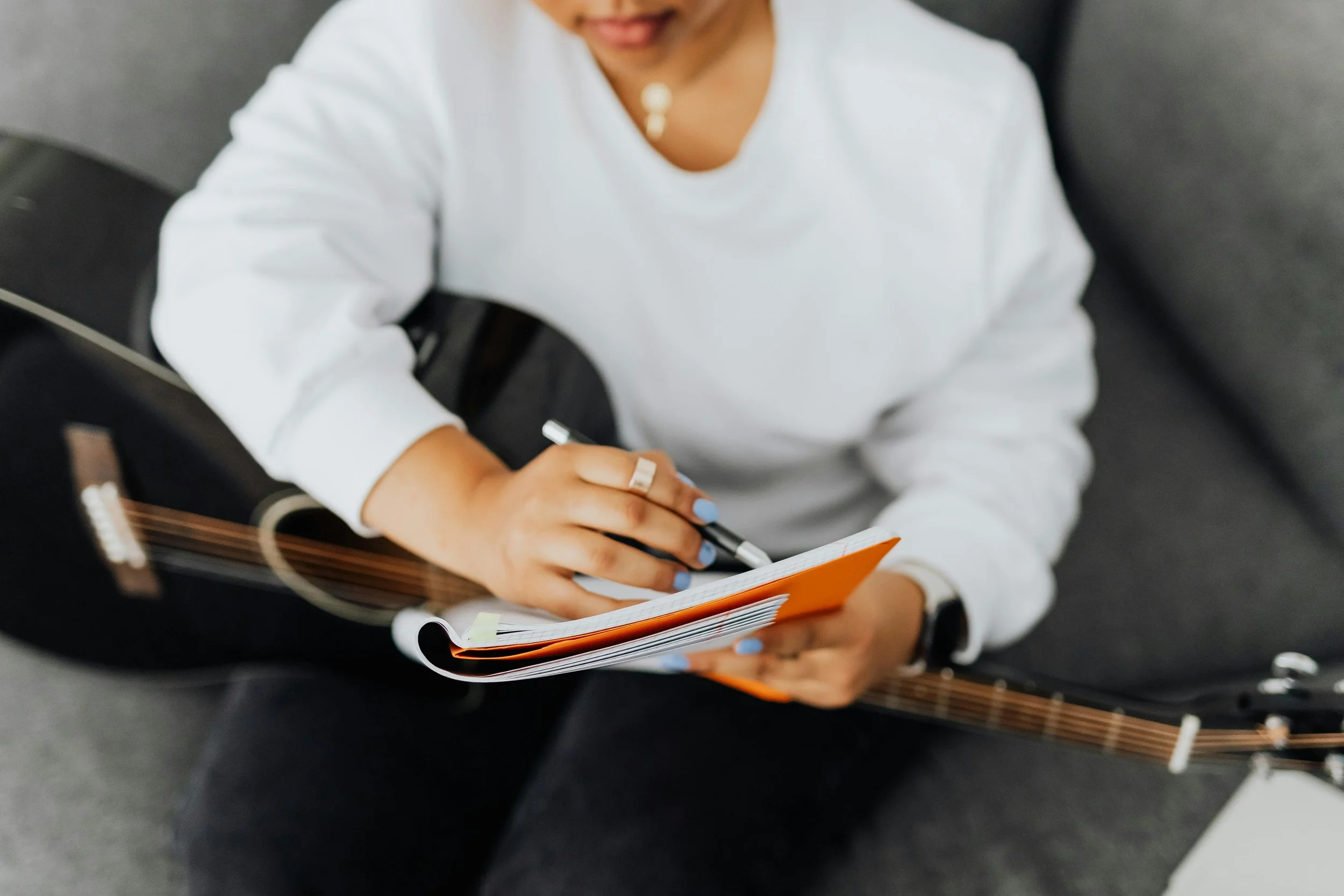 A person sitting on a gray couch writing in a notebook while holding an acoustic guitar.