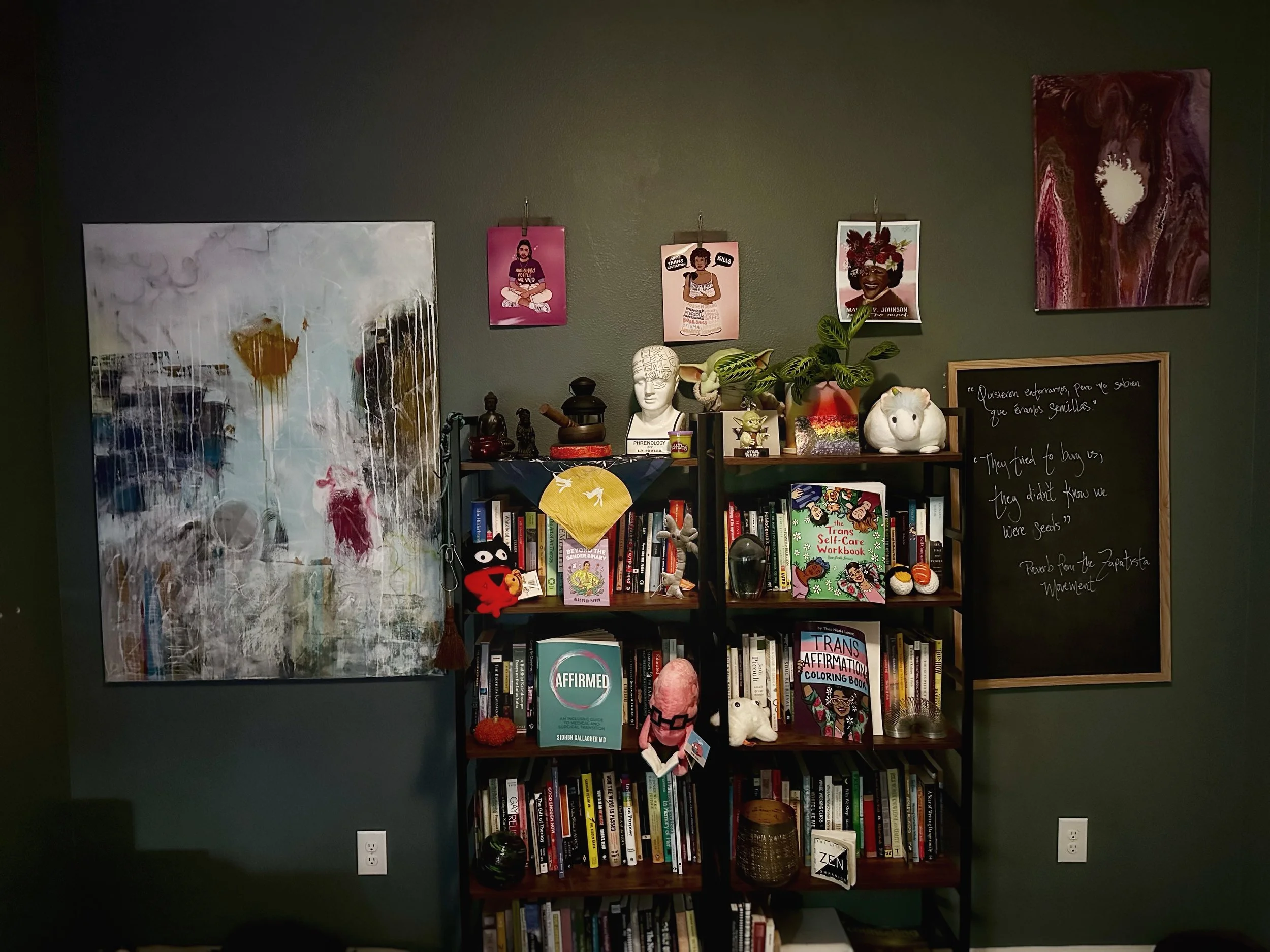 A bookshelf filled with books, decorations, and small figurines. Above the bookshelf are pictures of women, a stuffed animal, and plants. To the right of the shelf is a blackboard with writing, and to the left is an abstract painting on a green wall.