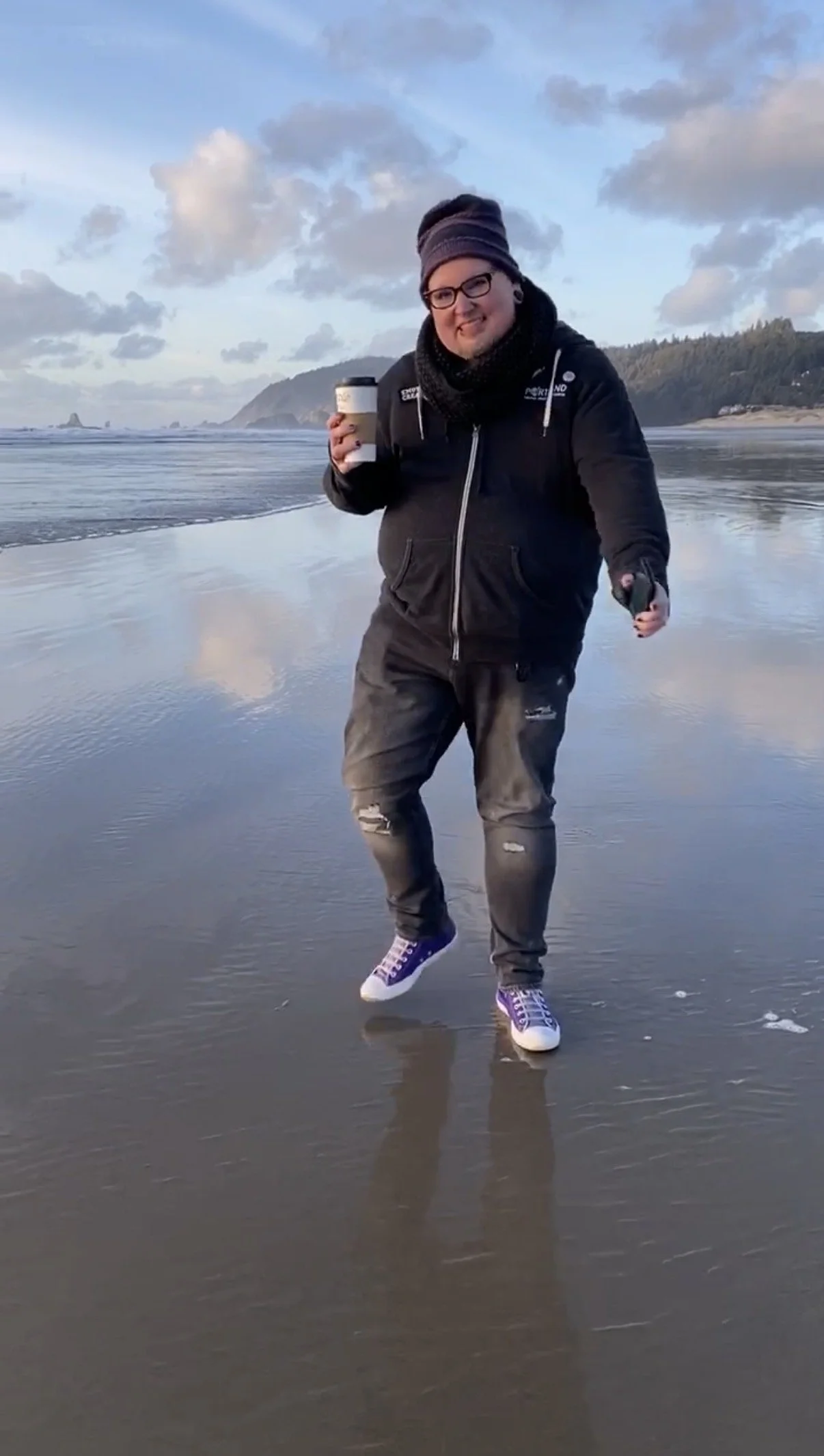genderqueer individual with glasses, wearing a beanie,holding a cup of chai at Cannon Beach