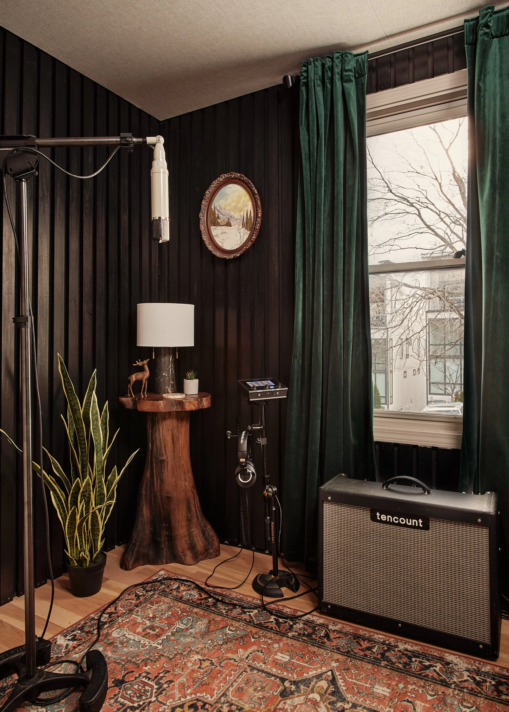 Recording studio corner with microphone, music stand, vinyl record player, amplifier, lamp, potted plant, and framed landscape picture on dark wall, green curtains, window, and patterned rug.