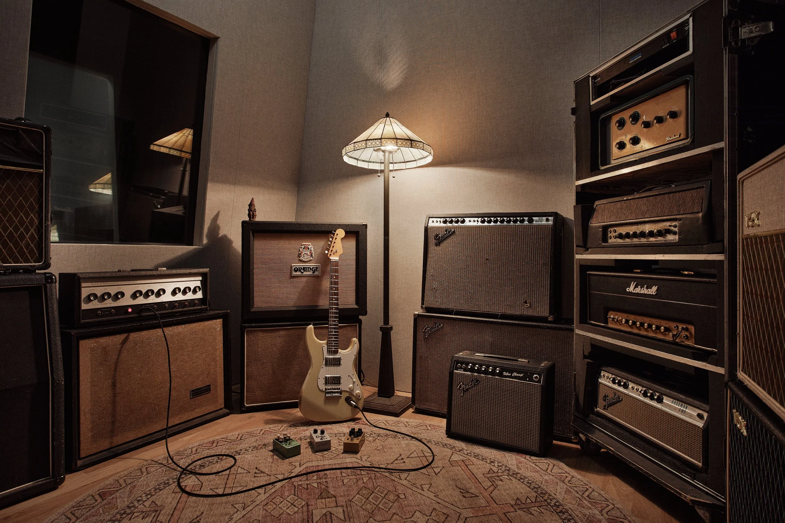 Music studio with guitar, vintage amplifiers, and pedals, lit by a floor lamp.