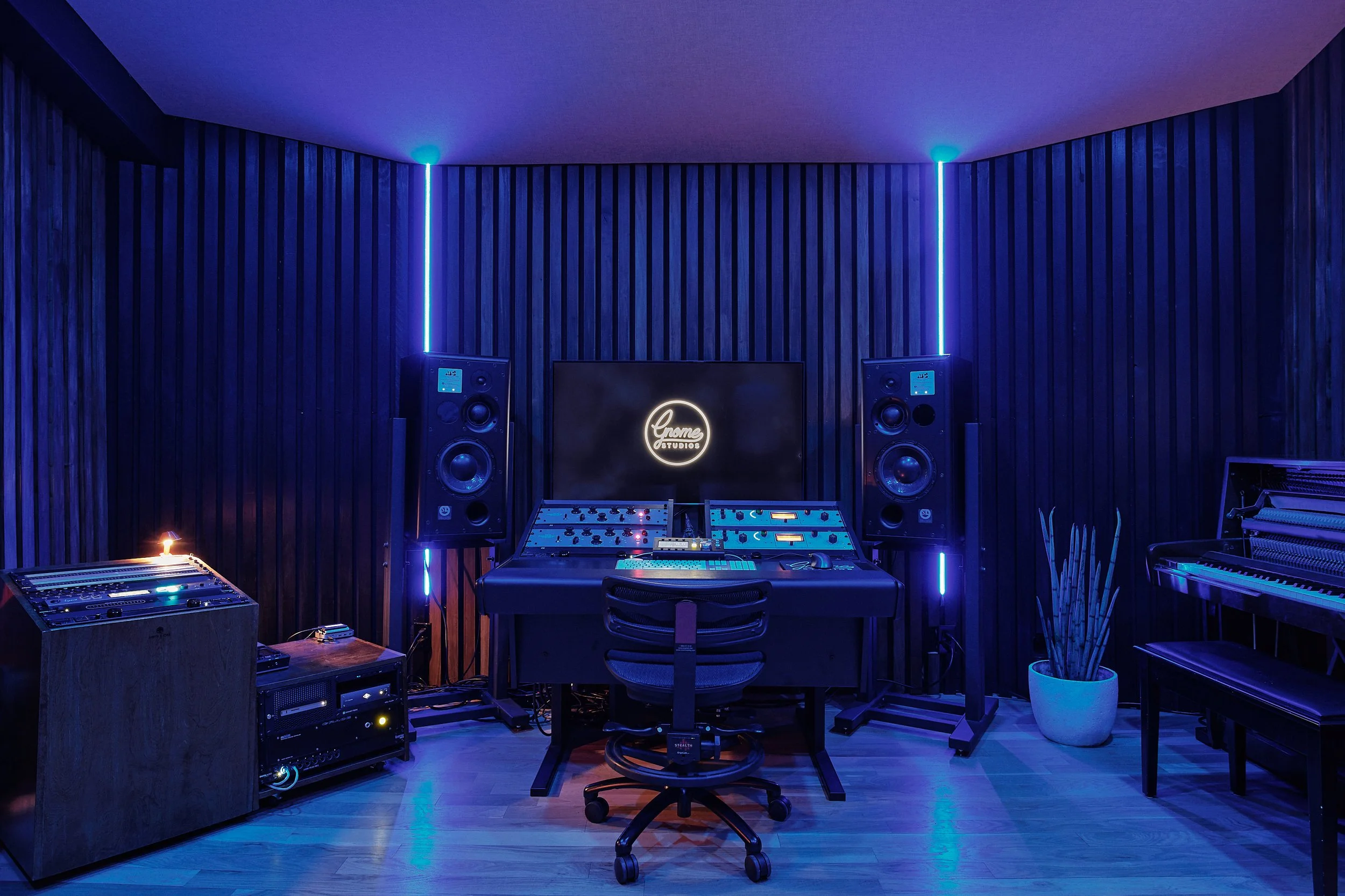 Recording studio with black walls, large speakers, mixing console, monitor screen displaying 'Gnome Studios', and various musical equipment, illuminated with blue lighting.
