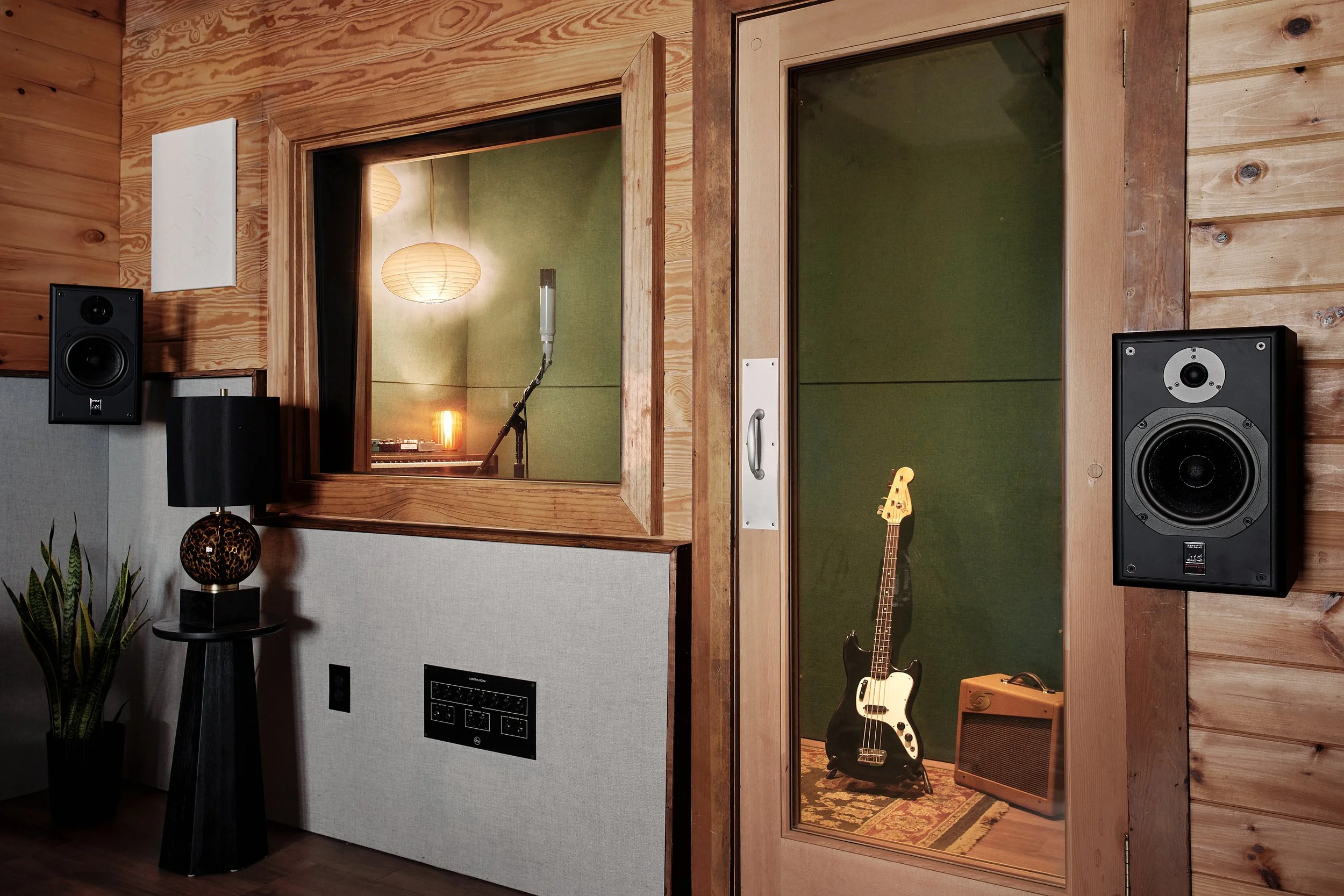 Sound recording studio with wooden walls, two speakers mounted on the wall, a window looking into the vocal booth with microphone, guitar, and amplifier inside.