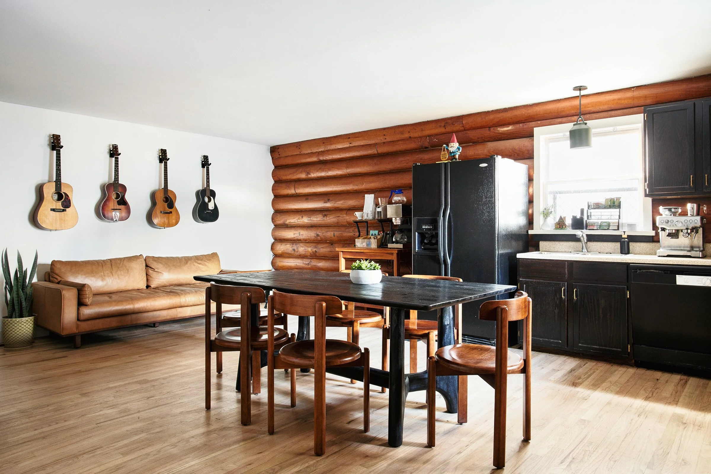 A cozy living room and kitchen area with four guitars hanging on a white wall, a tan leather sofa, a black dining table with six wooden chairs, and a black refrigerator. The kitchen has black cabinets, a window above the sink, and a small coffee make