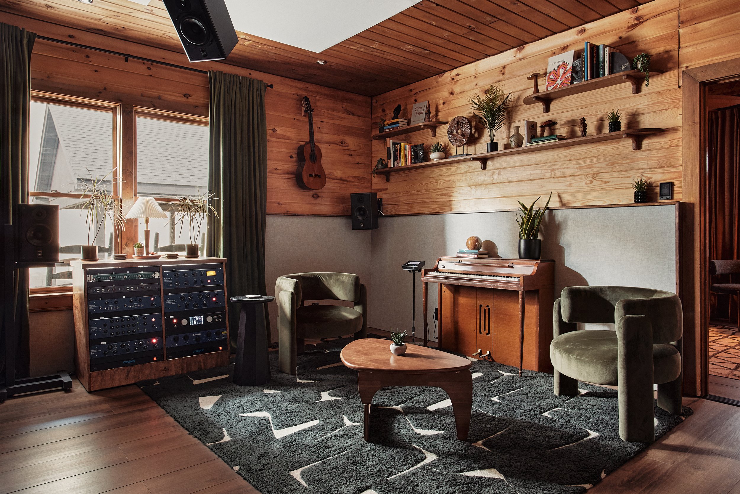 A cozy home recording studio with wooden walls and ceiling, featuring a large window with curtains, a speaker system, a guitar hung on the wall, a vinyl record, potted plants, a vintage piano, and comfortable green chairs.
