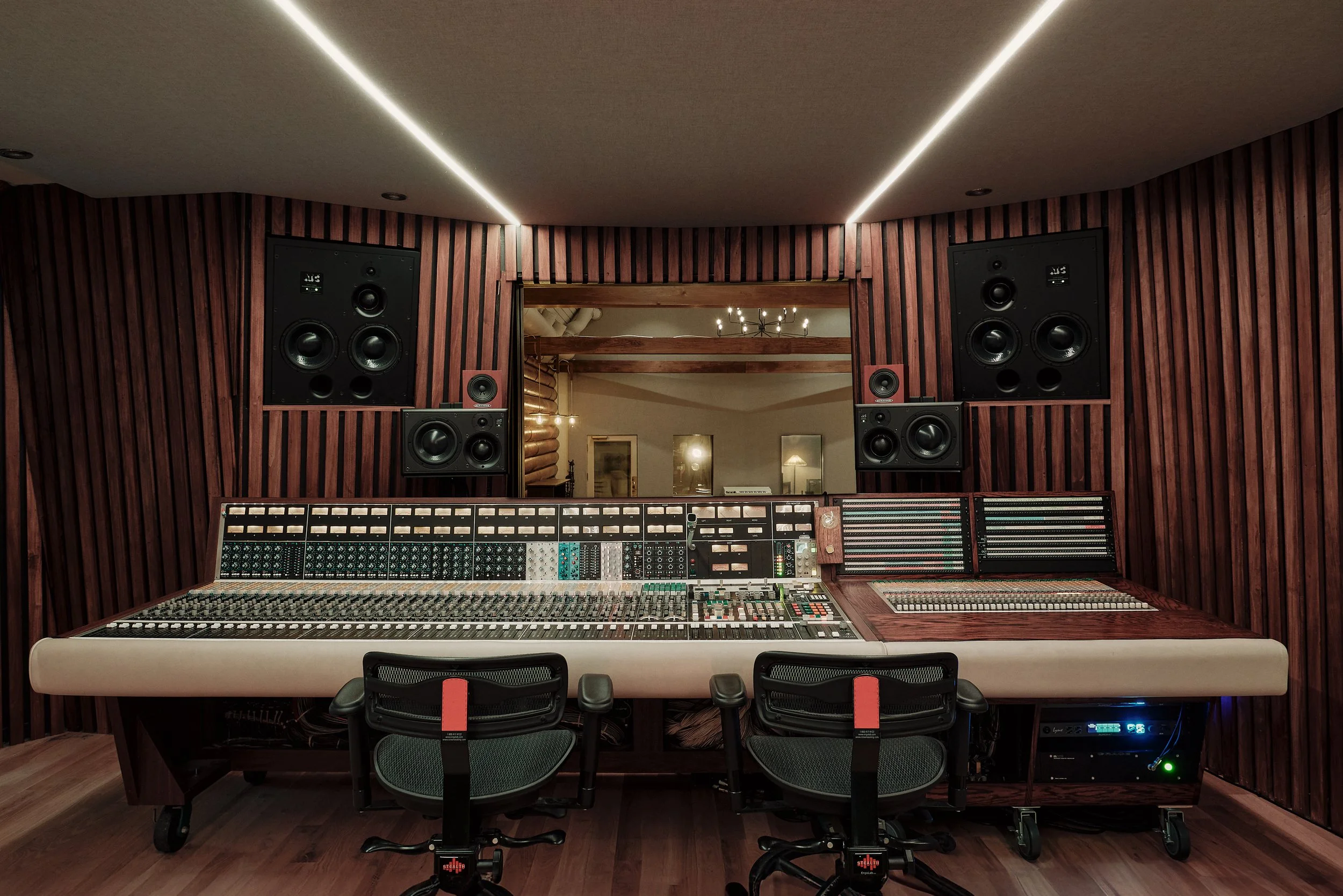 Recording studio with a large mixing console, two chairs, and multiple speakers on wood-paneled walls with reading window to another room.