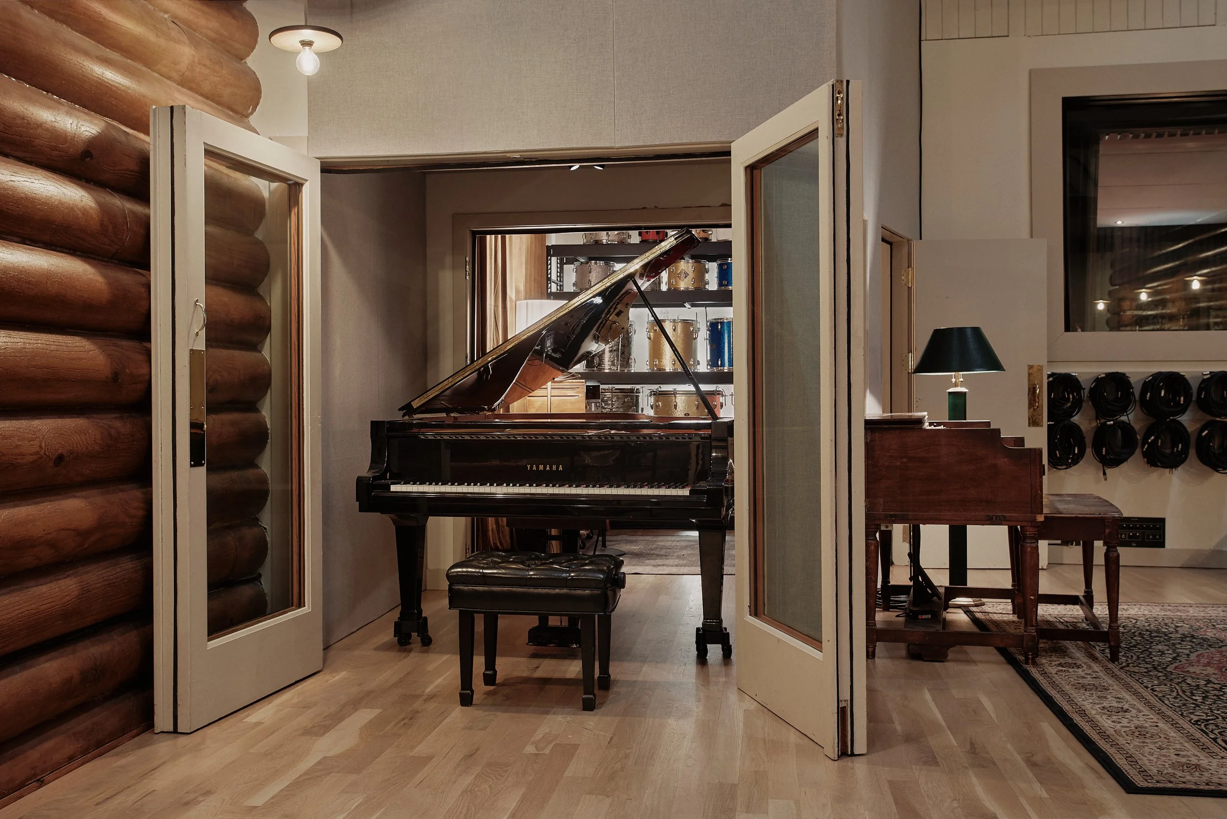 A black Yamaha grand piano with a matching bench in a music studio room, surrounded by wood and beige walls, with a black lamp and headphones on shelves.