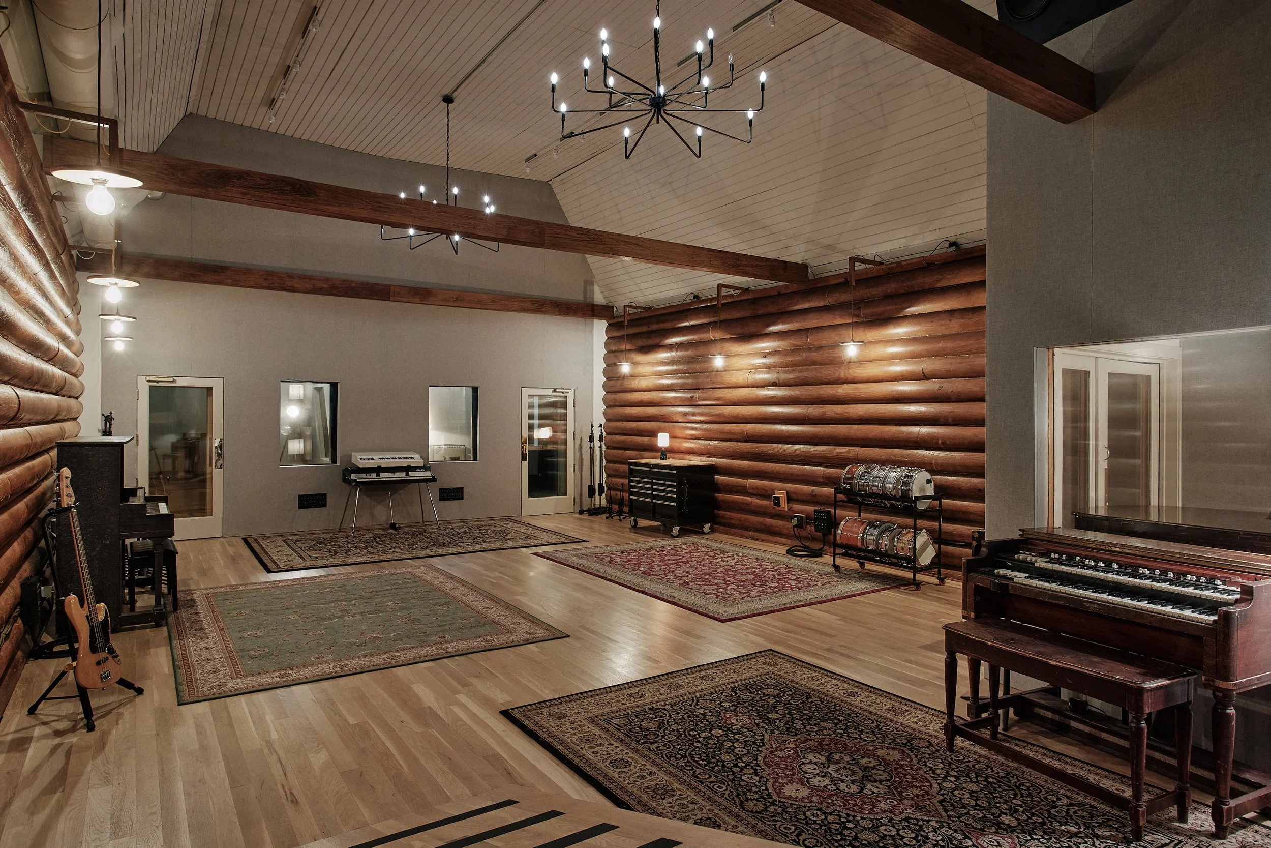 Recording studio with wooden log walls, hardwood floors, multiple vintage-style rugs, pianos, guitars, and music equipment, decorated with modern and rustic lighting fixtures.