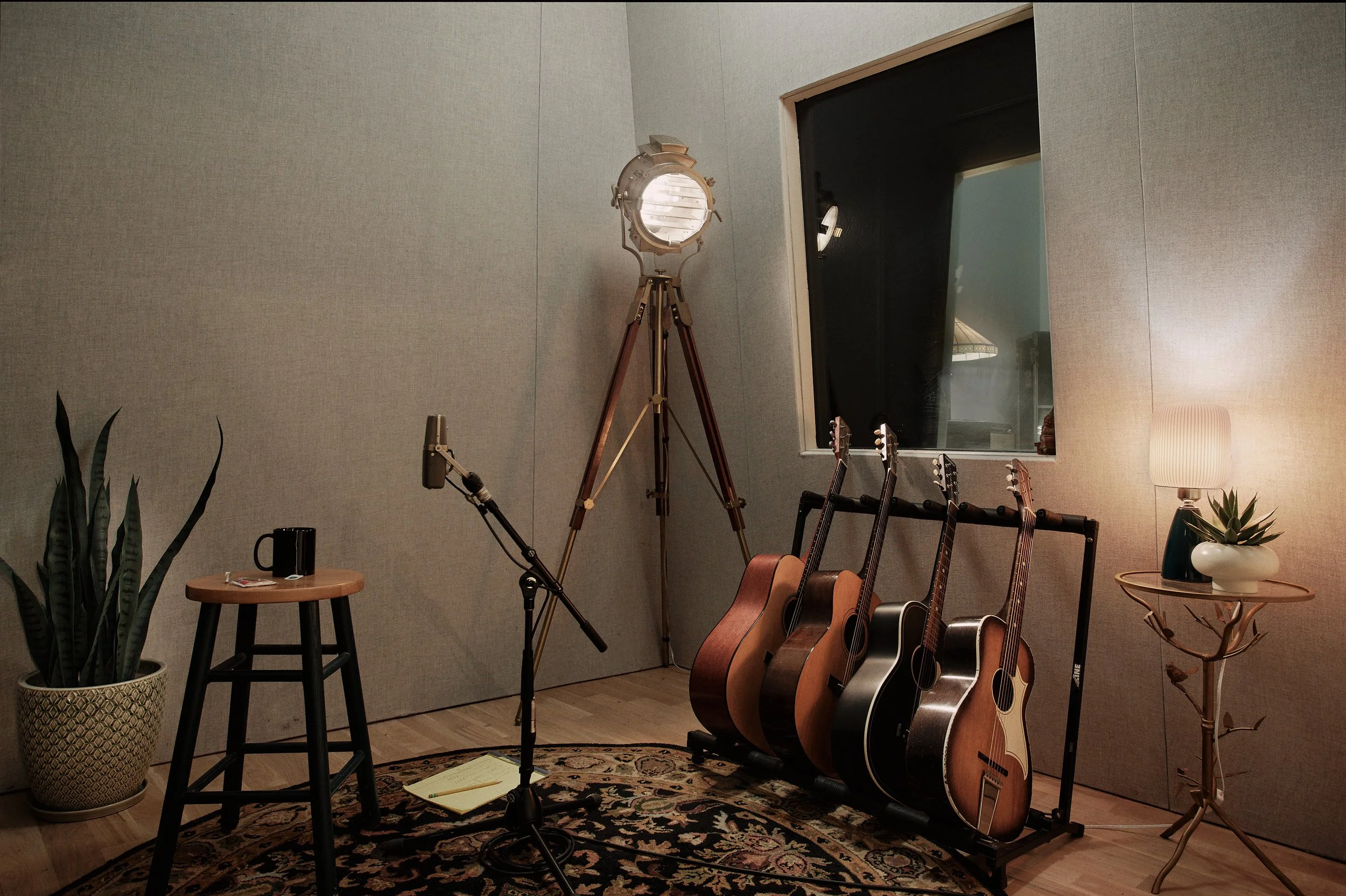 A music studio room with a tripod floor lamp, a plant in a decorative pot, a small side table with a mug and some keys, four acoustic guitars on a rack, a microphone on a stand, and a table with a lamp and a potted plant. There is a window on the wal