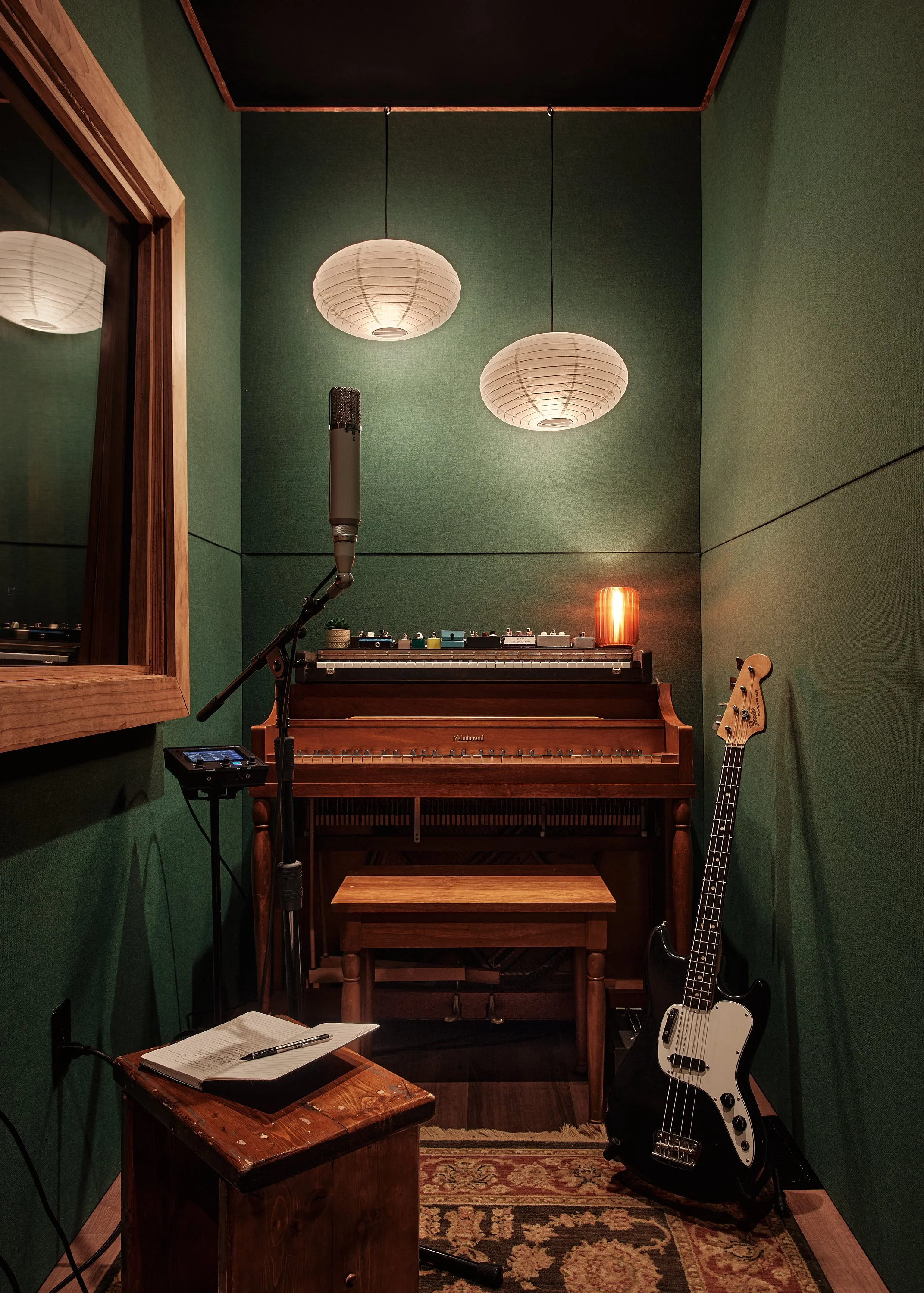 Soundproof music recording studio with green walls, a wooden upright piano, a bass guitar, a microphone on a stand, a music stand, a small notebook, and hanging paper lanterns.