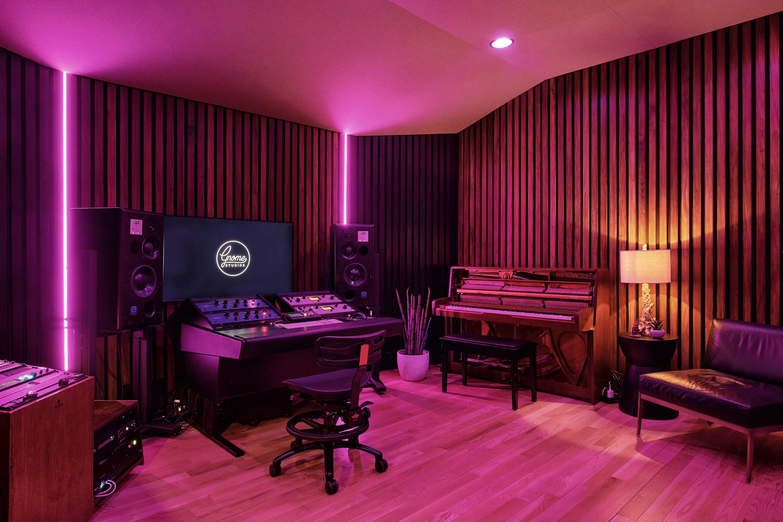 A recording studio with wooden acoustic panels, pink and purple LED lighting, a computer monitor displaying 'Gnome Studios,' large black studio speakers, a mixing console, a music keyboard, a black office chair, a small round table with a lamp and pl