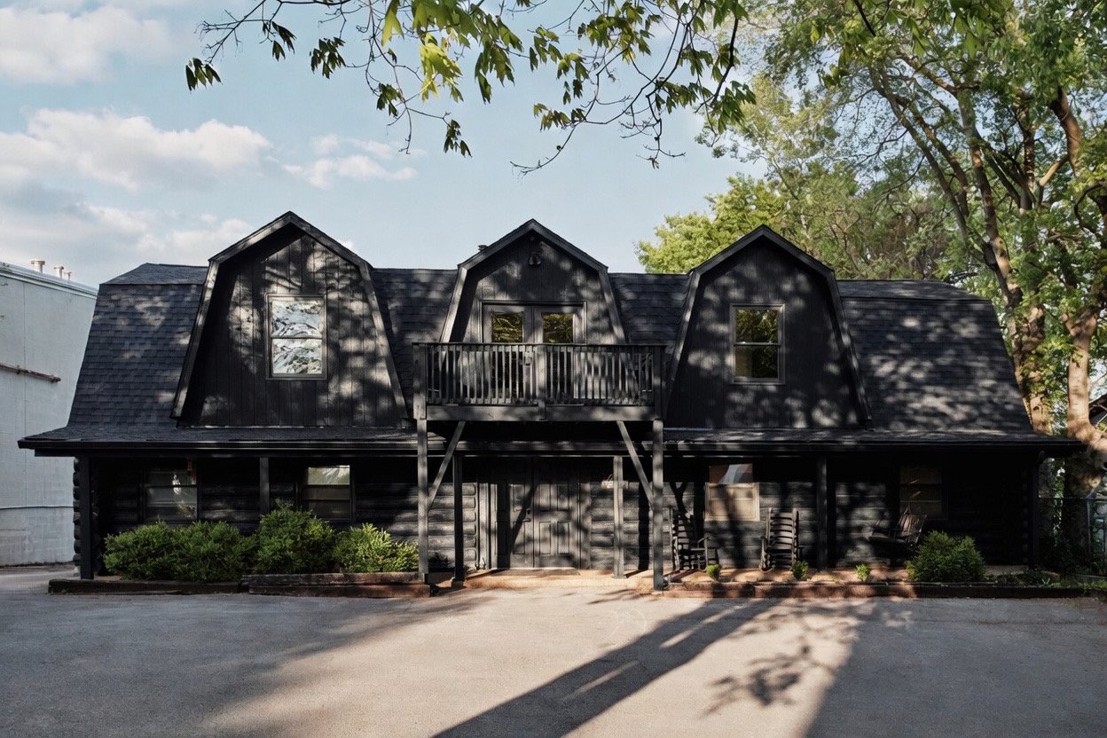 A black, two-story house with a steep roof and dormer windows, shadowed by trees, with a small balcony and a parking area in front.