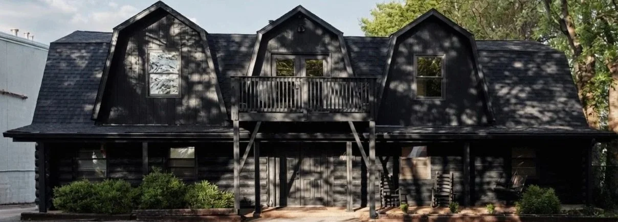 A two-story black wooden house with a small central balcony, surrounded by trees and greenery, with outdoor chairs on the patio.