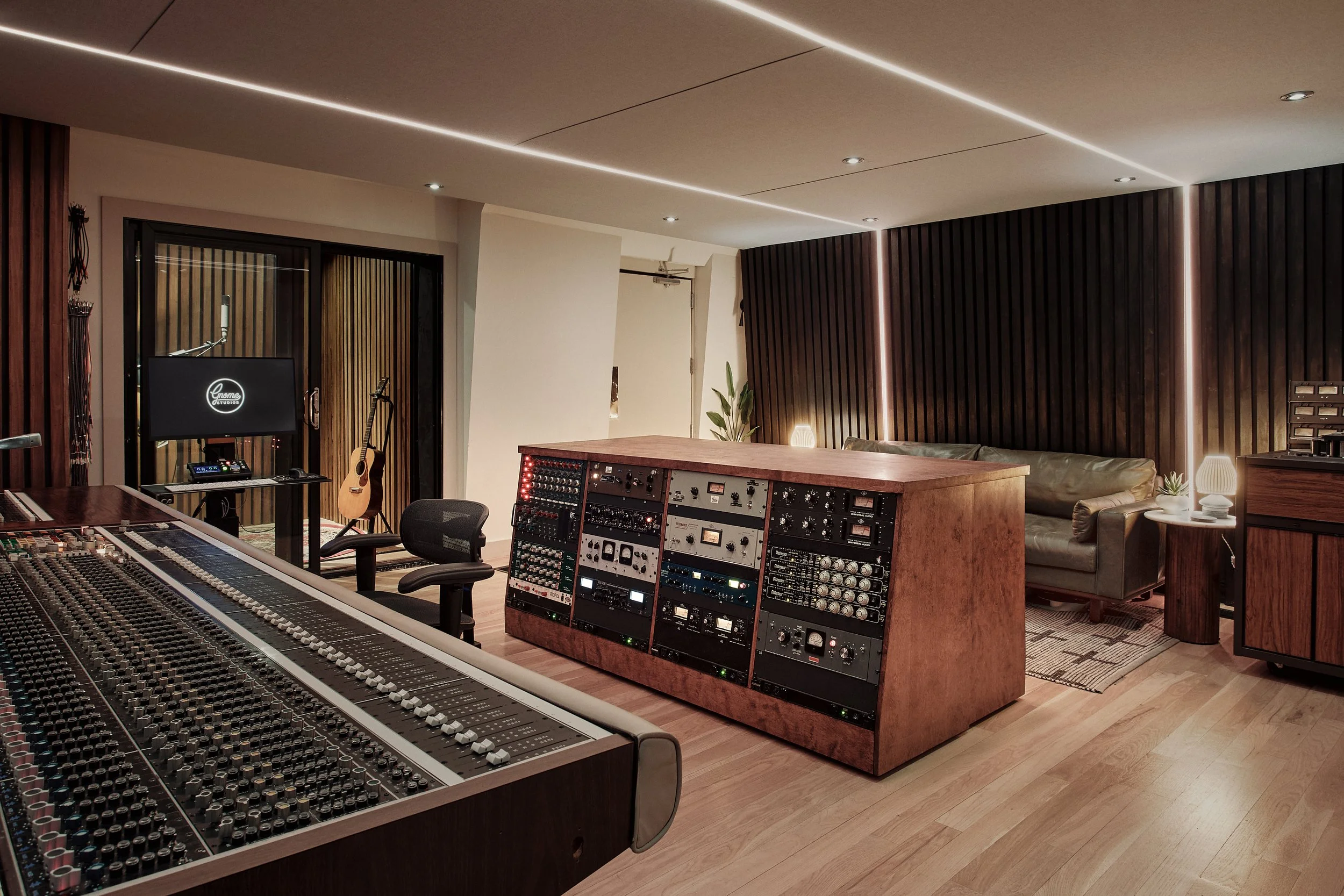 A professional recording studio with a mixing console, sound equipment, and a cozy sitting area with lamps, plants, and a guitar.