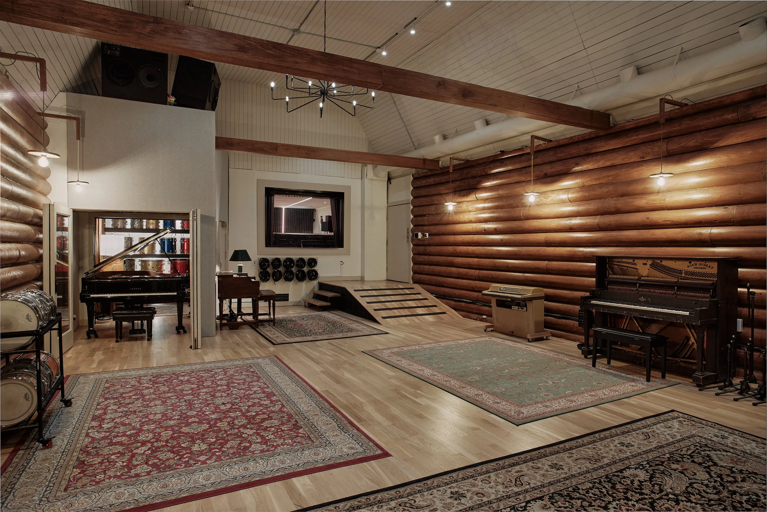 A music rehearsal room with wooden floors and log walls, featuring a grand piano, a vintage upright piano, drums, and various rugs.