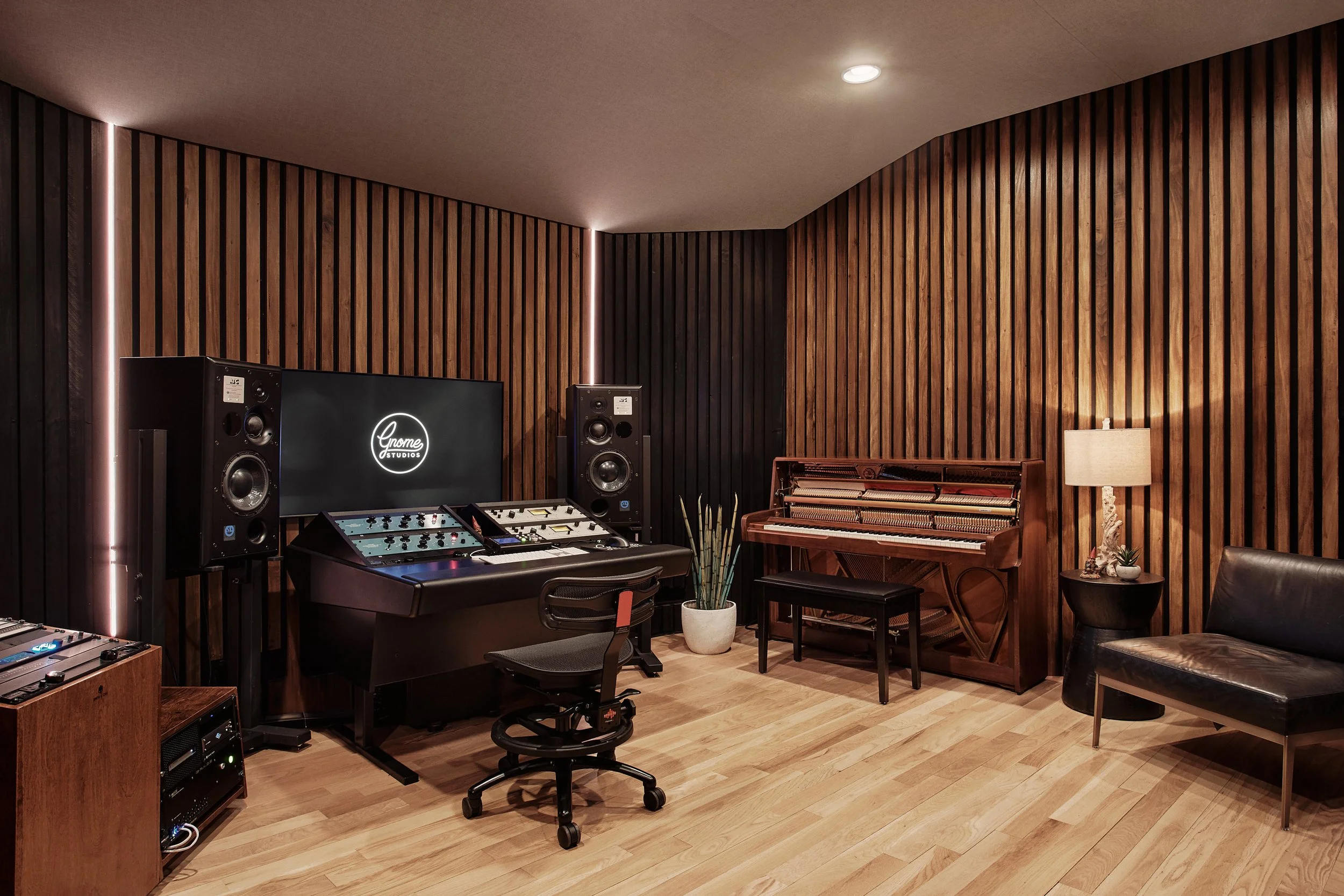 Recording studio with wooden panel walls, a large monitor displaying 'Gnome Studios', audio equipment, a piano, a black leather chair, a floor lamp, and a potted plant.
