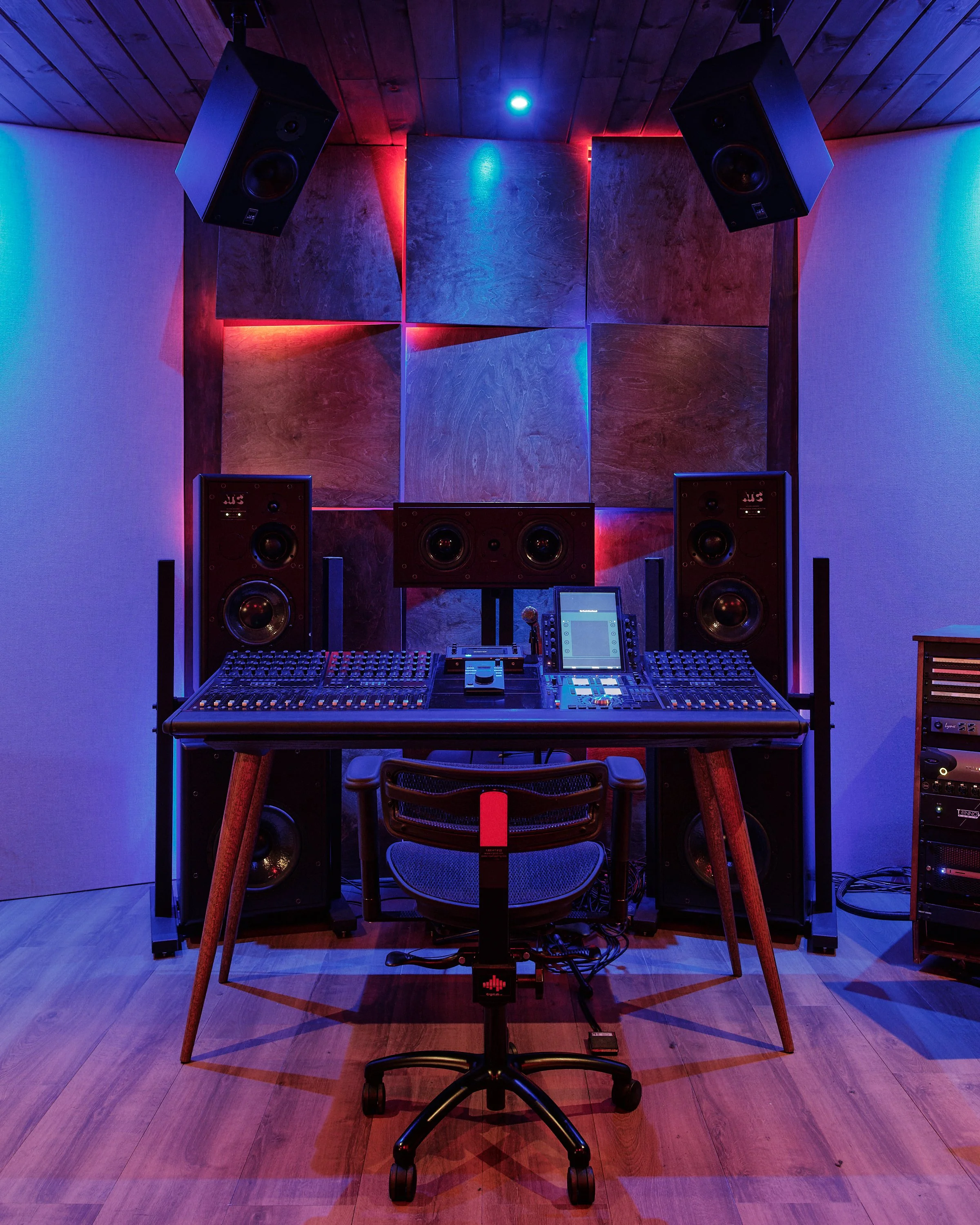 Audio recording studio with mixing consoles, large speakers, and sound-absorbing wooden panel background illuminated by blue and red LED lights.