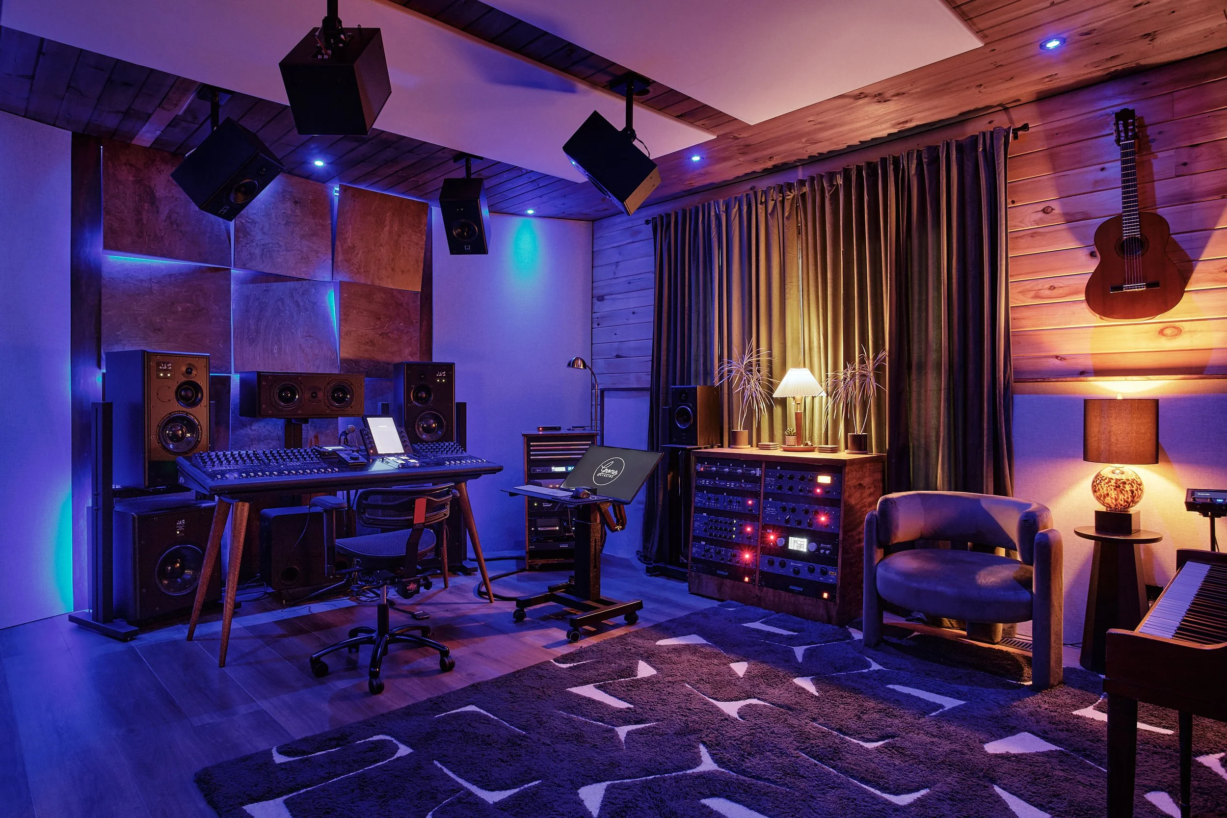 A recording studio with wooden walls, various electronic music equipment, large speakers, a mixing console, a computer, a lamp, plants, a guitar hanging on the wall, also featuring a cozy chair, a side lamp, a carpet, and curtains.