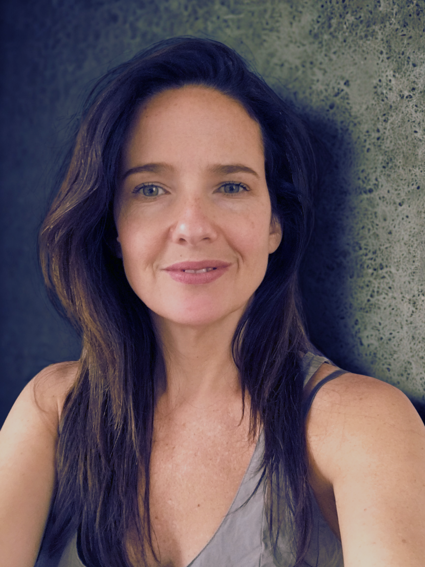 A woman with blue eyes and long dark hair is smiling at the camera, posing in front of a textured wall.
