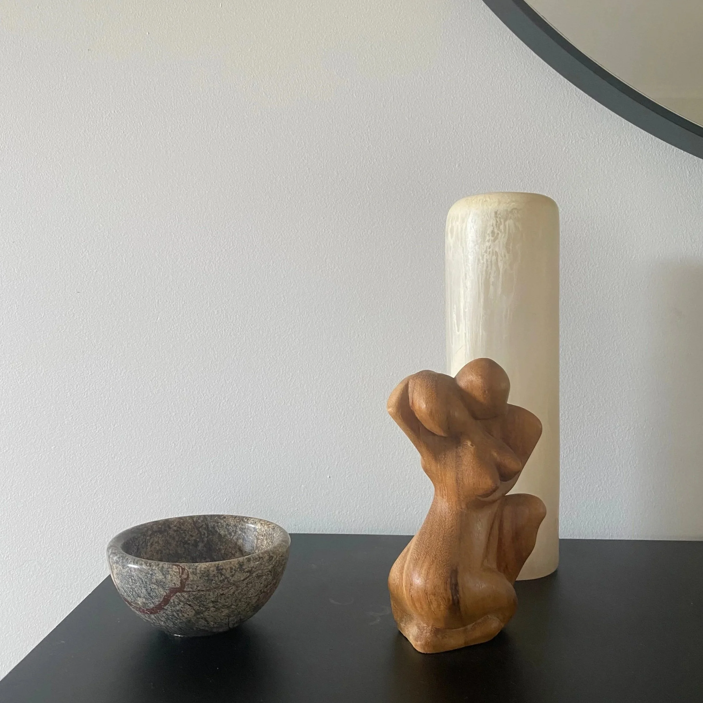 Decorative objects on a black table including a white ceramic vase, a wooden abstract sculpture, and a small stone bowl.
