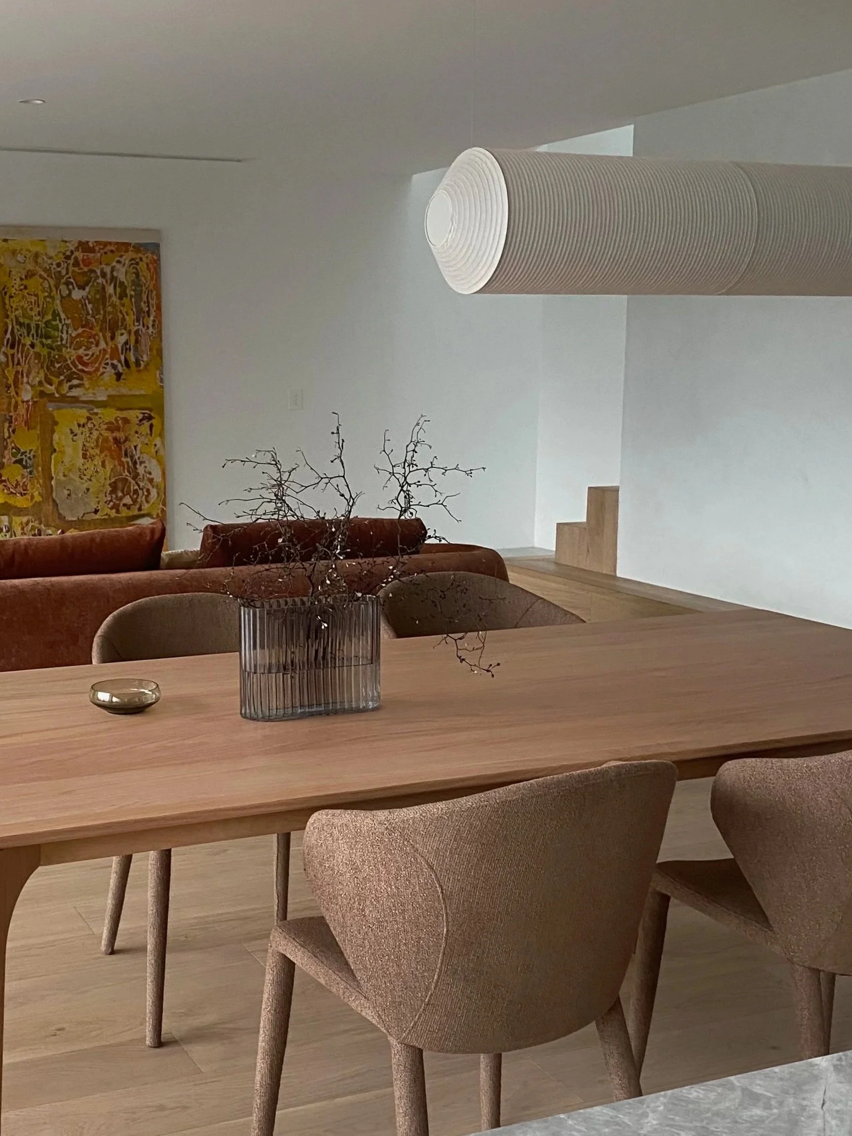 A modern dining room with a wooden table, beige upholstered chairs, a glass vase with dry branches, and abstract artwork on the wall in the background.