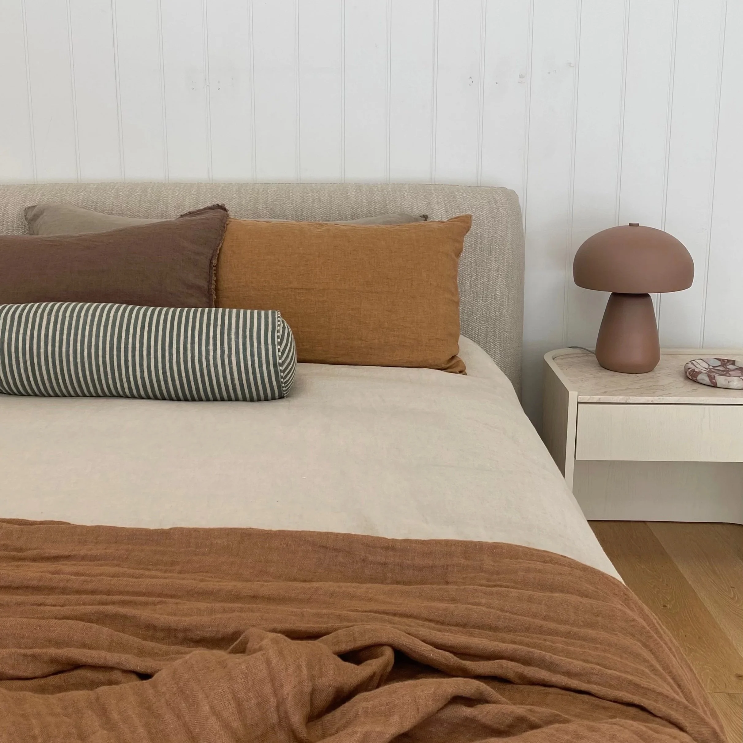 A bed with beige bedding, brown and striped pillows, a white wooden nightstand with a mushroom-shaped pink lamp, and a decorative stone dish in a minimalist bedroom with white paneled walls and hardwood floors.