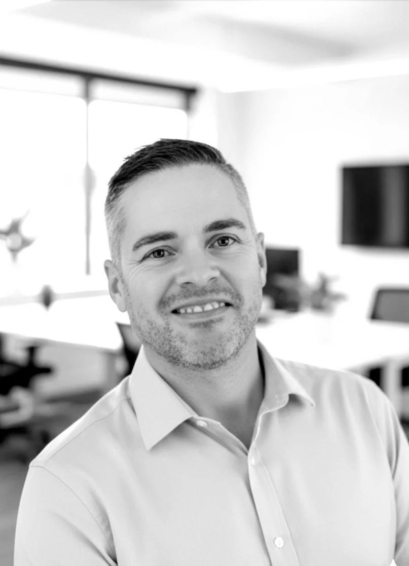 A smiling man with short hair and a beard wearing a light-colored collared shirt, in an office environment.