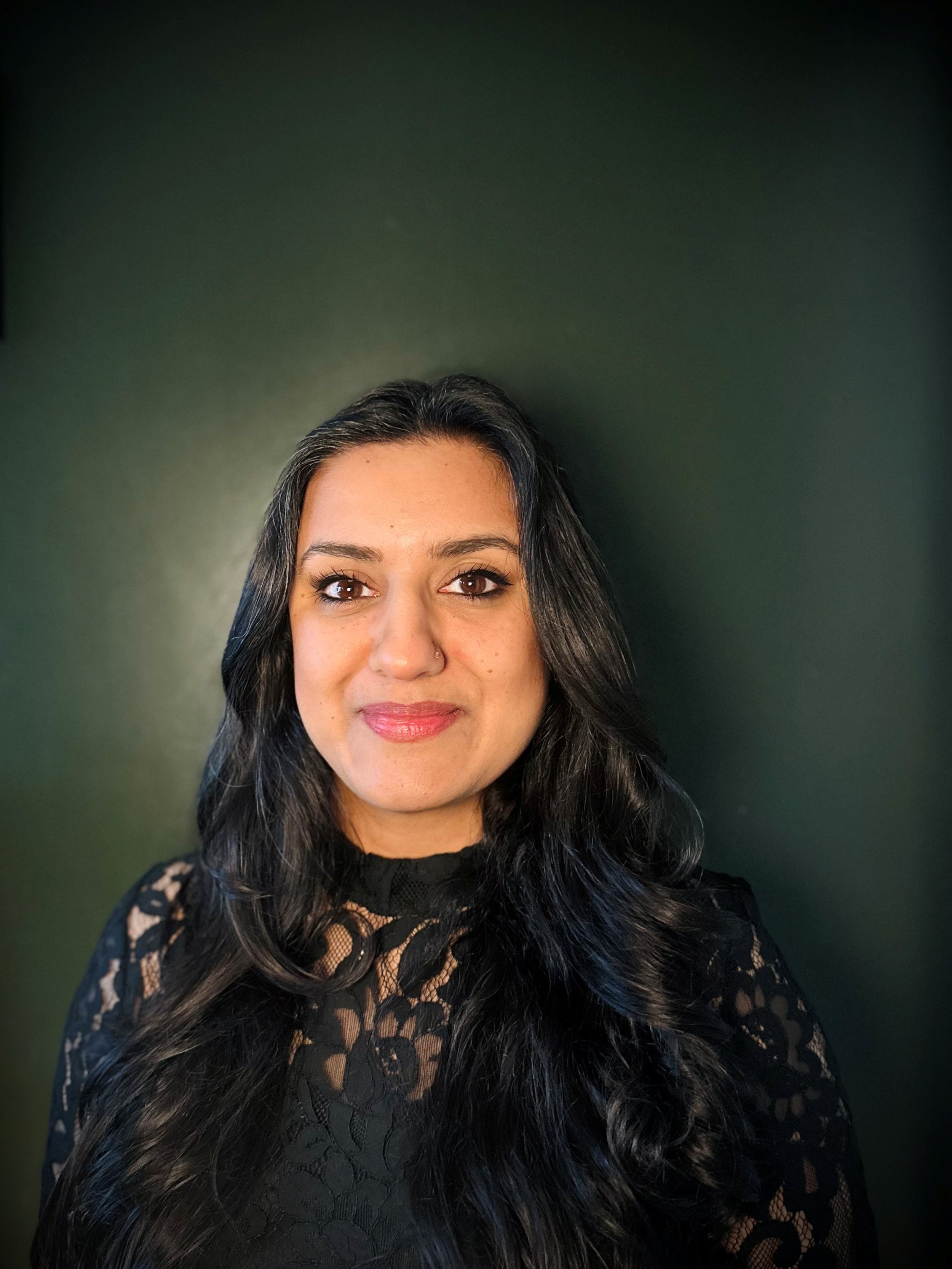 A woman with long, dark wavy hair wearing a black lace top, standing against a dark green background.