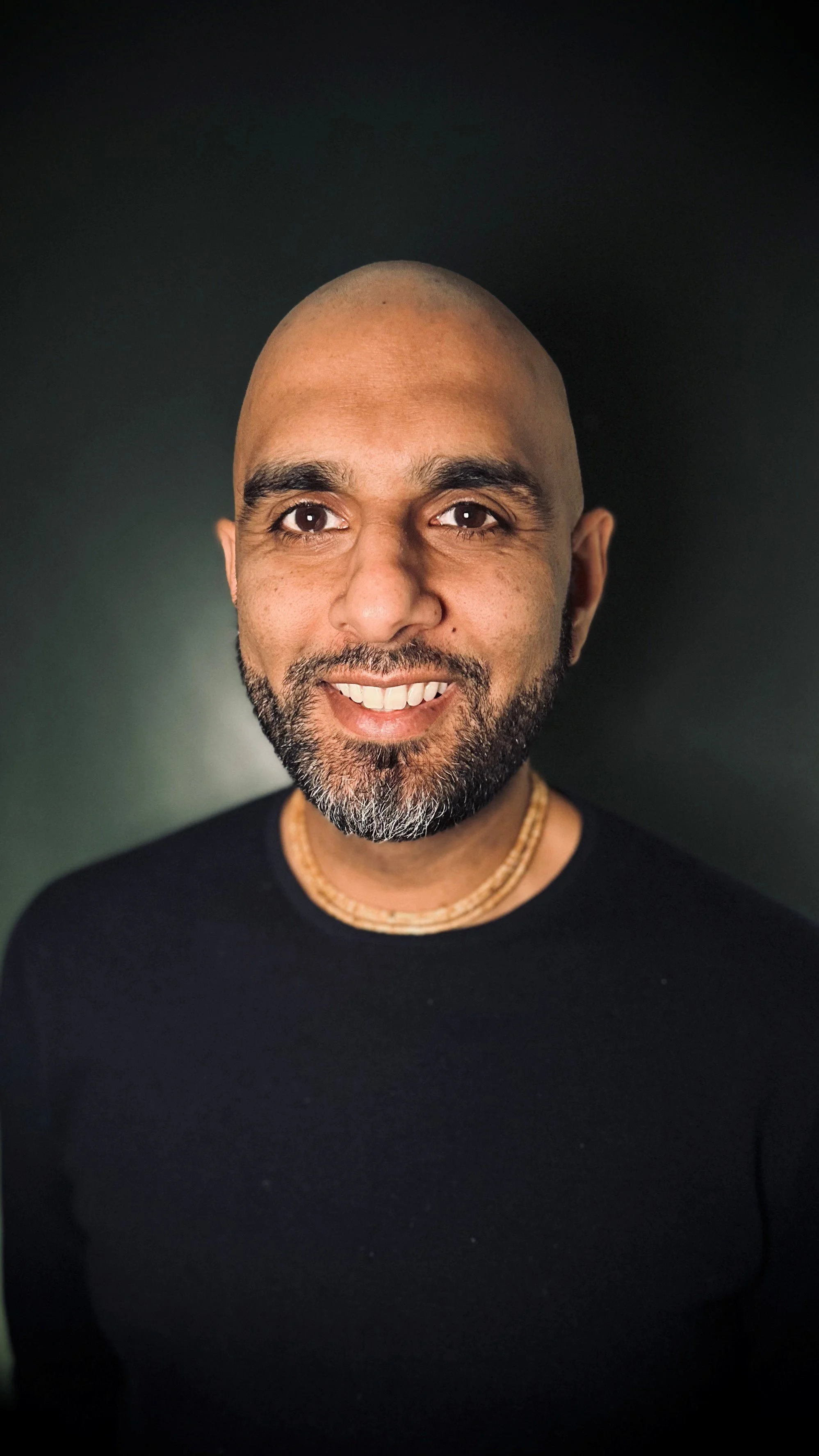 A smiling man with a shaved head and a beard, dressed in a black shirt and wearing a layered necklace, standing against a dark background.