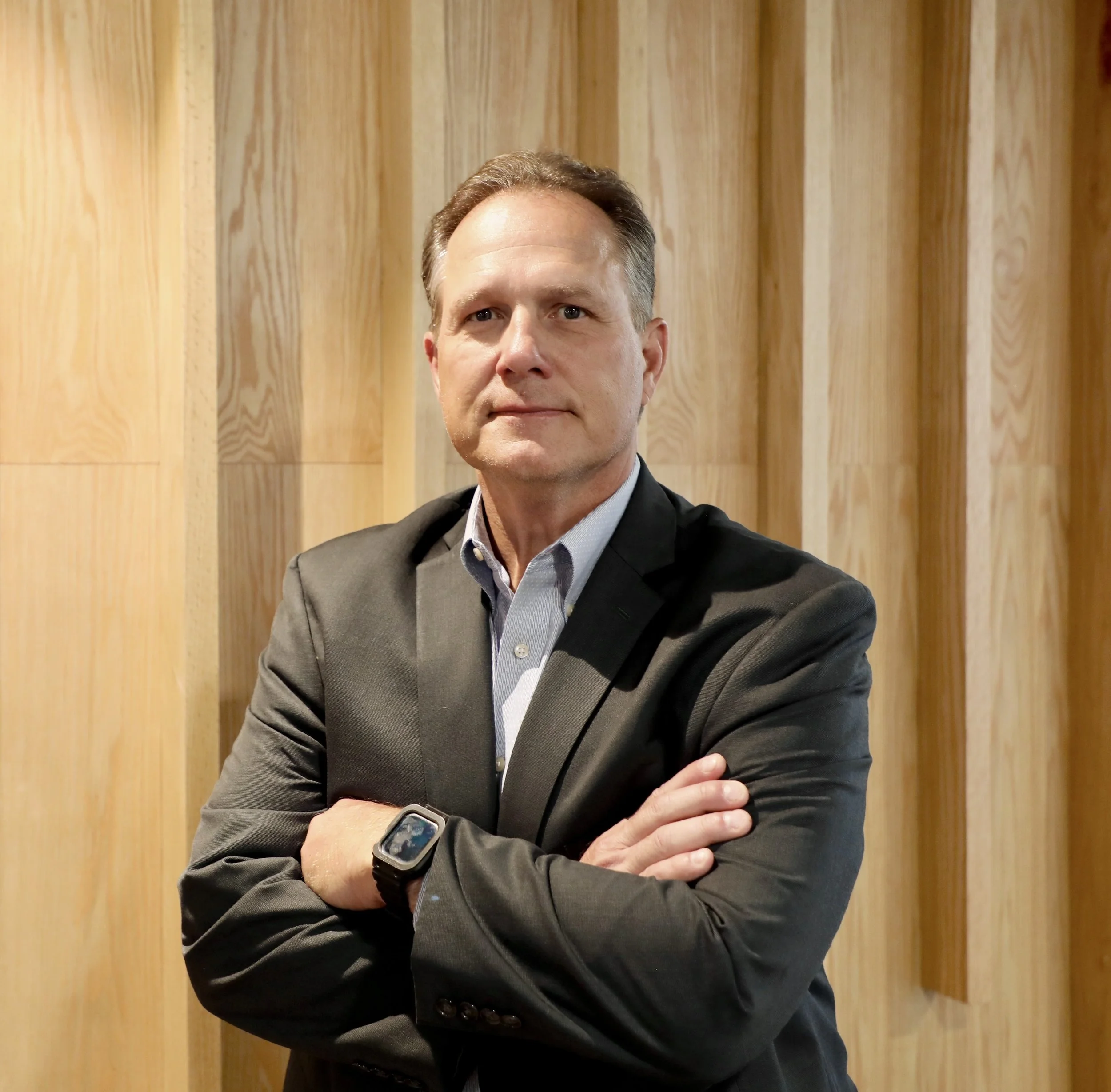 A man in a dark suit with arms crossed, standing in front of a wooden wall.